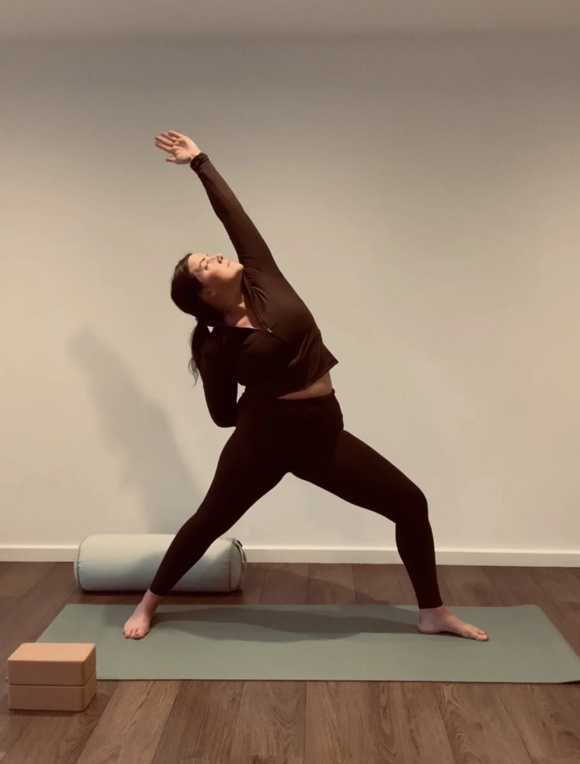 A woman practicing yoga indoors on a green mat, performing a side bend stretch with her left arm reaching upward and her right arm behind her back, wearing black leggings and a dark top.