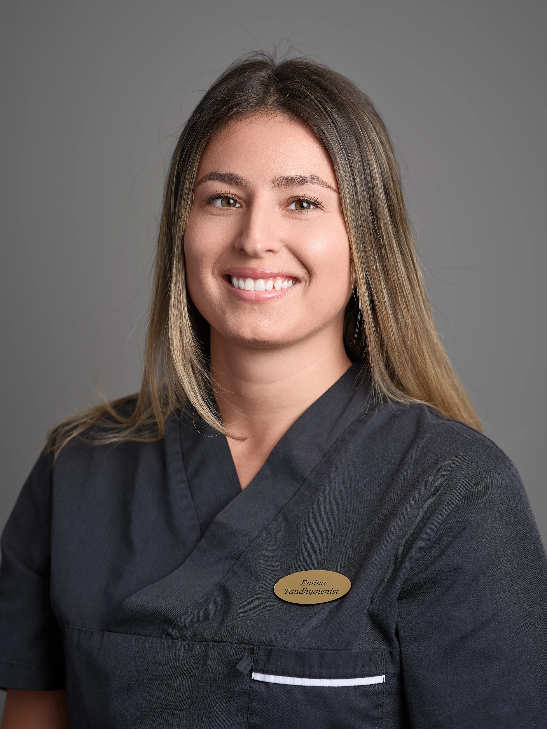 Porträtt av en ung kvinna i en mörk klinikkläder med namnskylt som säger 'Emina Tandhygienist', mot en grå bakgrund.