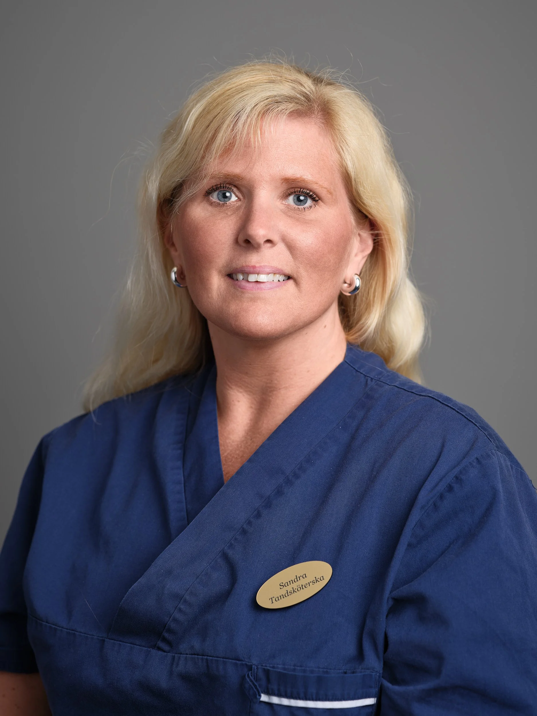 En kvinna med blont hår, blå ögon, och ett leende, bär en blå tandläkarsköterskeuniform med ett namnskylt som säger 'Sandra Tandhygienist'.