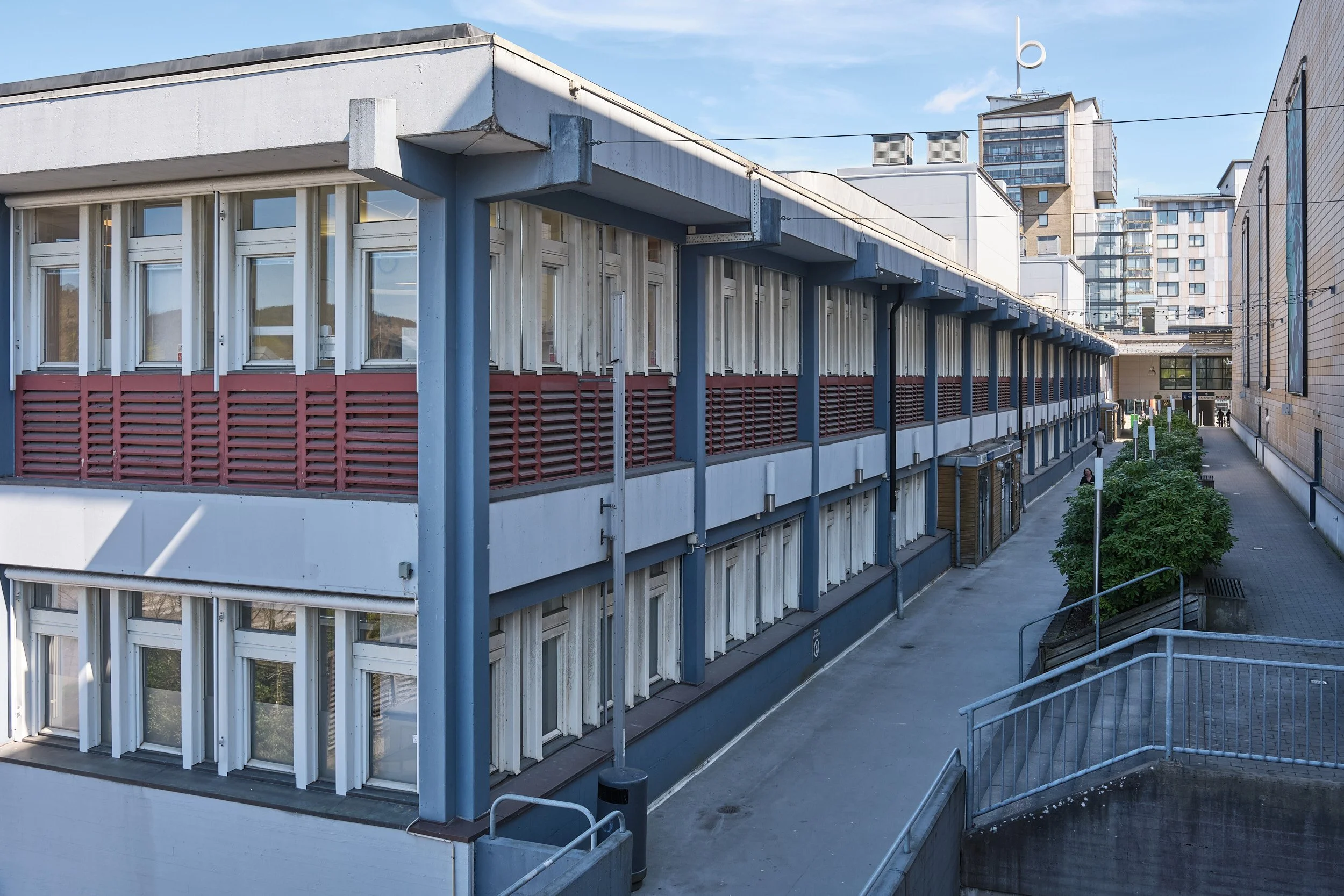 Utsikt över en modern byggnad med många fönster och blåa och röda detaljer, omgiven av höga flerbostadshus i city.