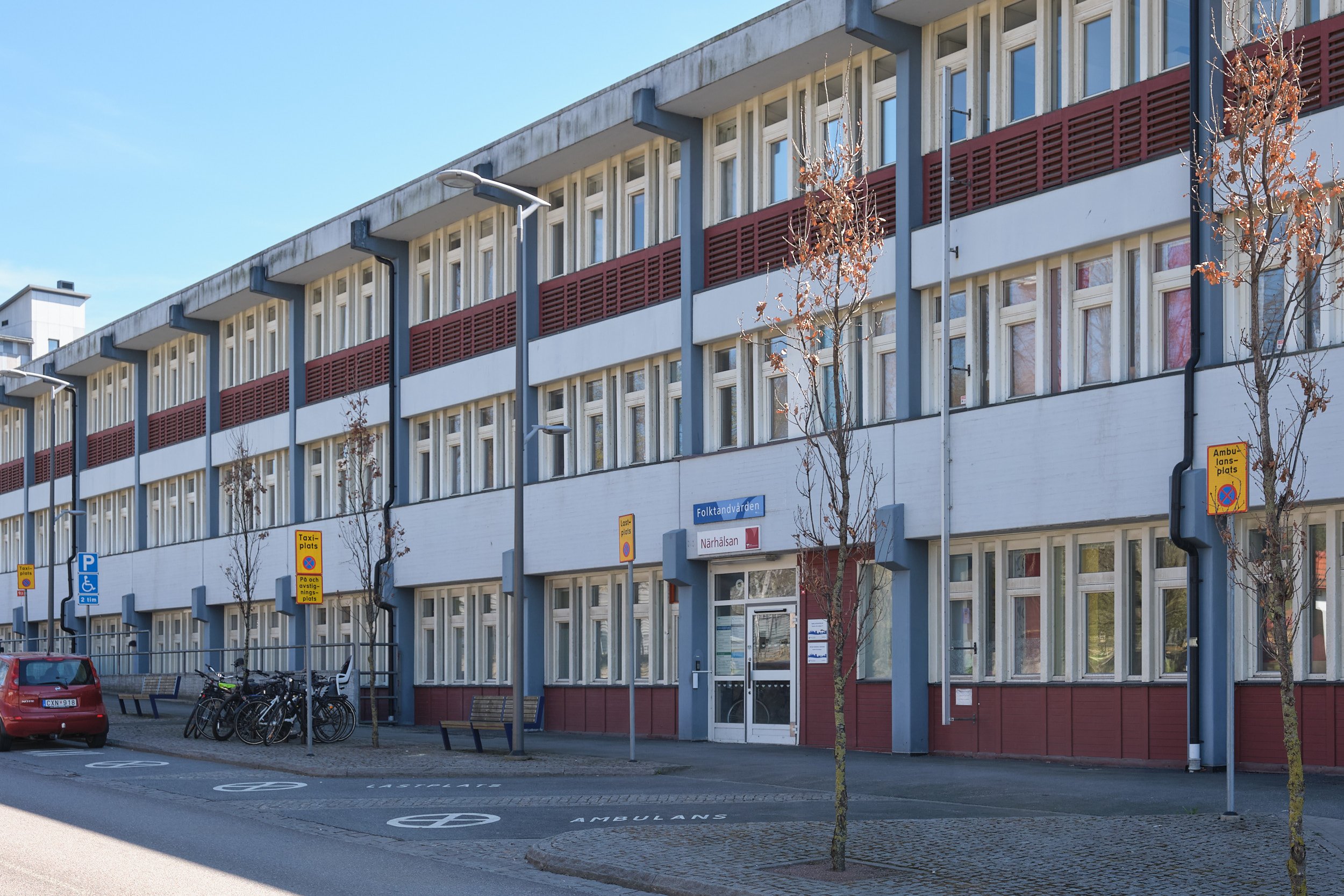 Bostadshus med många fönster, gatustolpar, träd, parkeringsplatser och skyltar, i ett stadsmiljö. Solig dag.