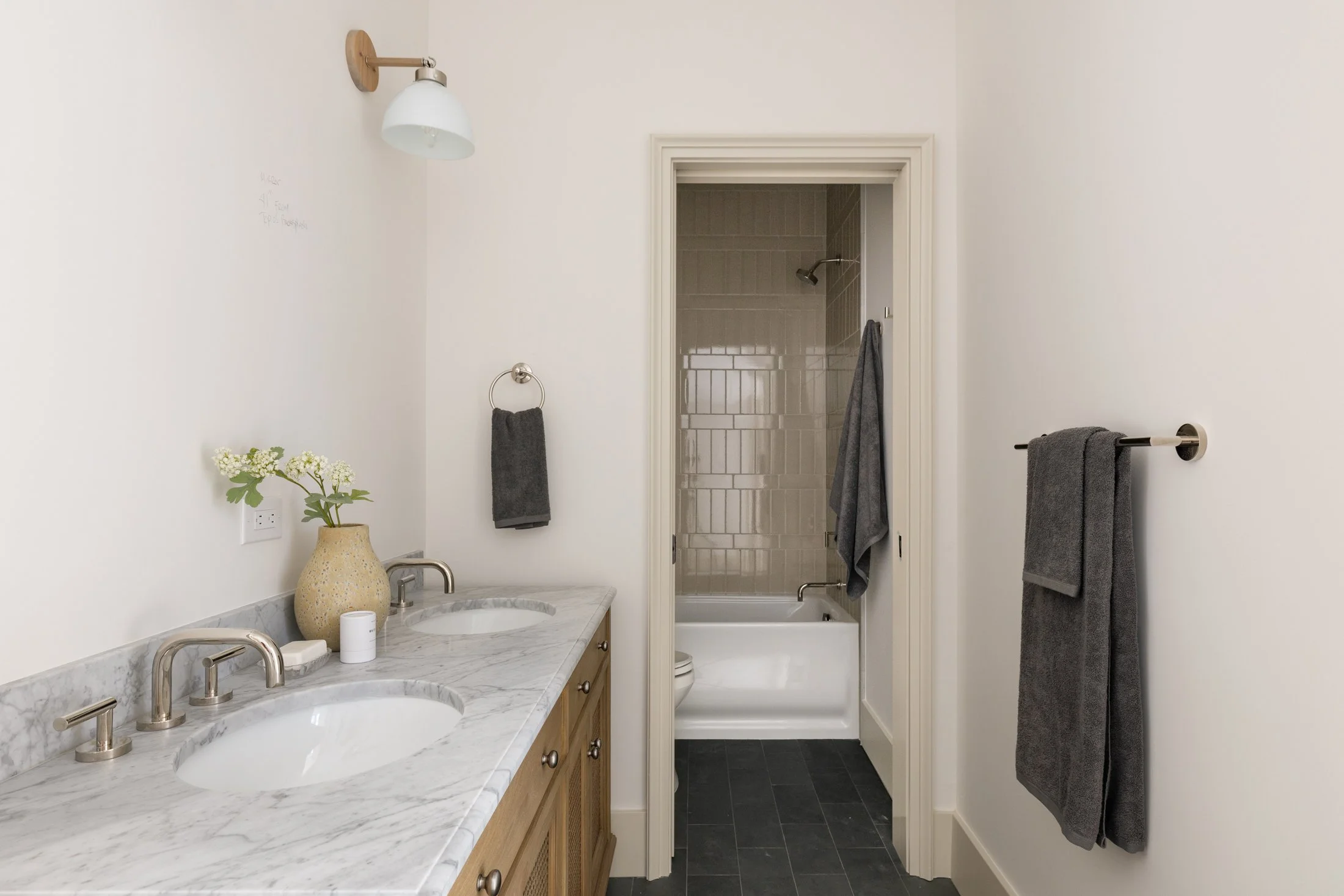 A bathroom with a dual-sink marble vanity, gray towels, a vase with white flowers, a doorway to a bathtub and shower, and a black tile floor.