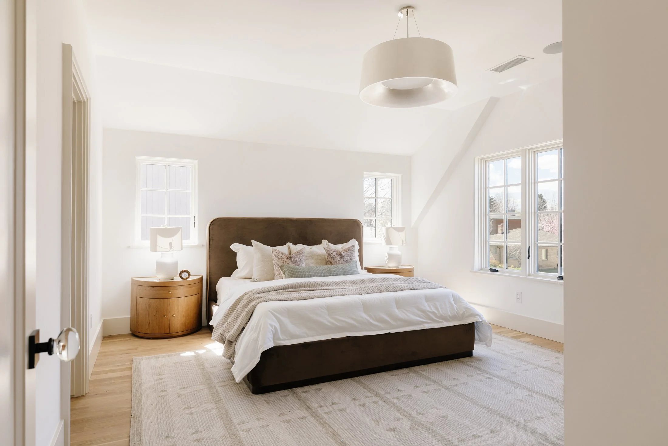 Bright bedroom with white walls, large windows, a bed with white bedding and multiple pillows, wooden nightstands with lamps, and a light-colored rug.