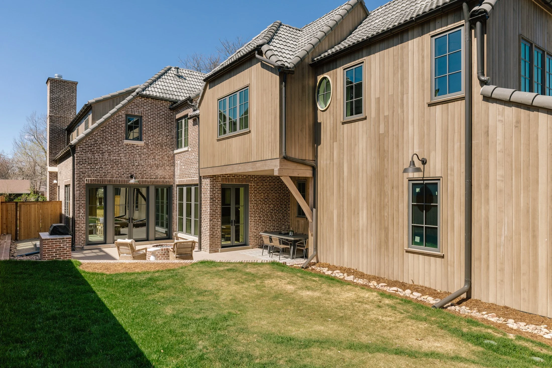 Backyard of a modern two-story house with brick and wood exterior, green lawn, patio furniture, and large windows.
