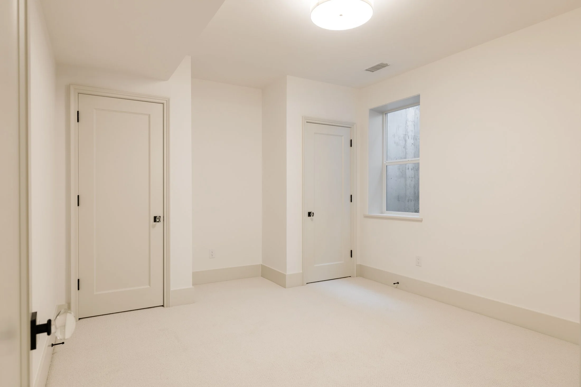 An empty, white-walled room with beige carpet, two closed doors, a window with condensation, a ceiling light, and an air vent.