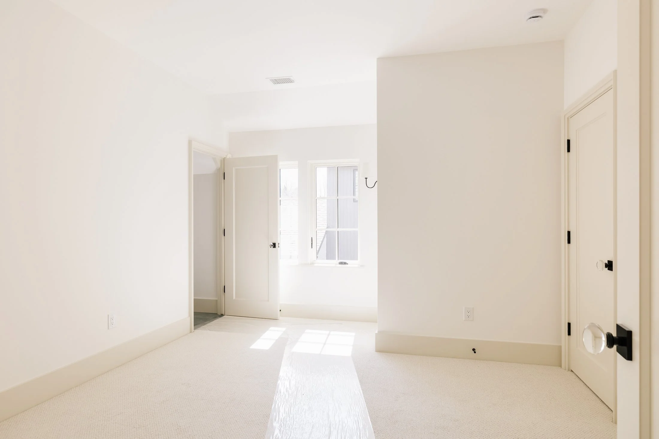 Bright, empty white room with a window, two open doors, white walls, beige carpet, and black door hardware.