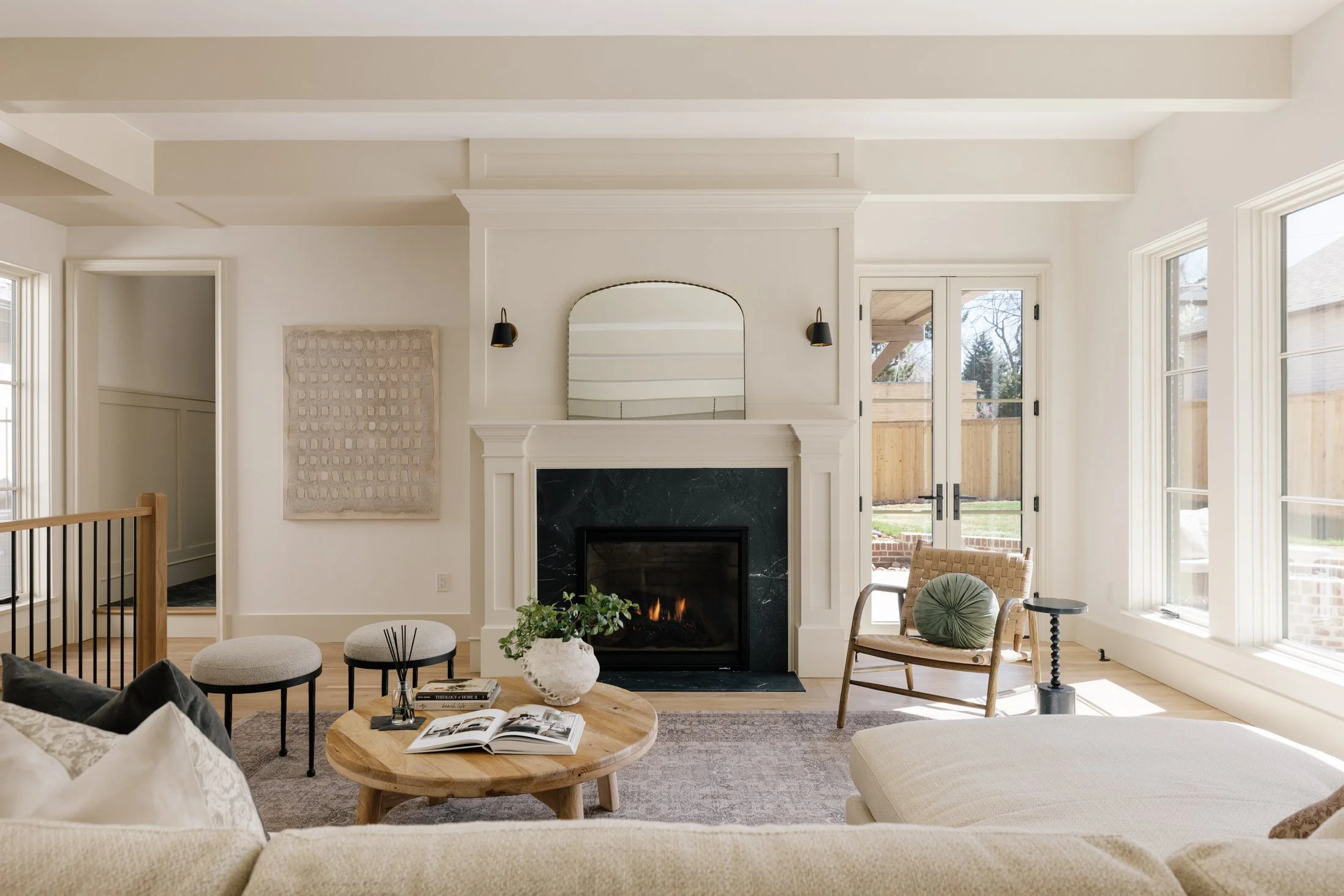 Bright living room with white walls, a fireplace with black marble surround, and a mirror above. Large windows and glass doors let in natural light, showing a fenced backyard. The room has a beige sofa, wooden coffee table, and two small stools. Deco