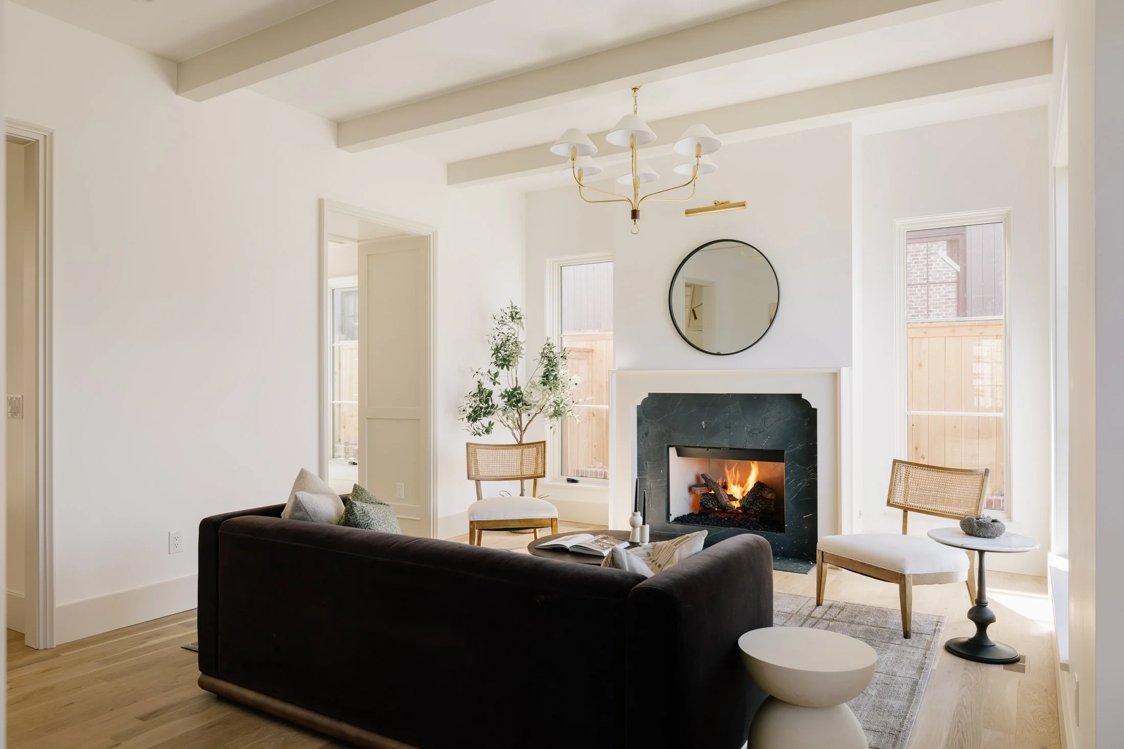 Living room with a black sofa, two white chairs, a fireplace with a fire, round mirror on the wall, and a chandelier hanging from the ceiling.