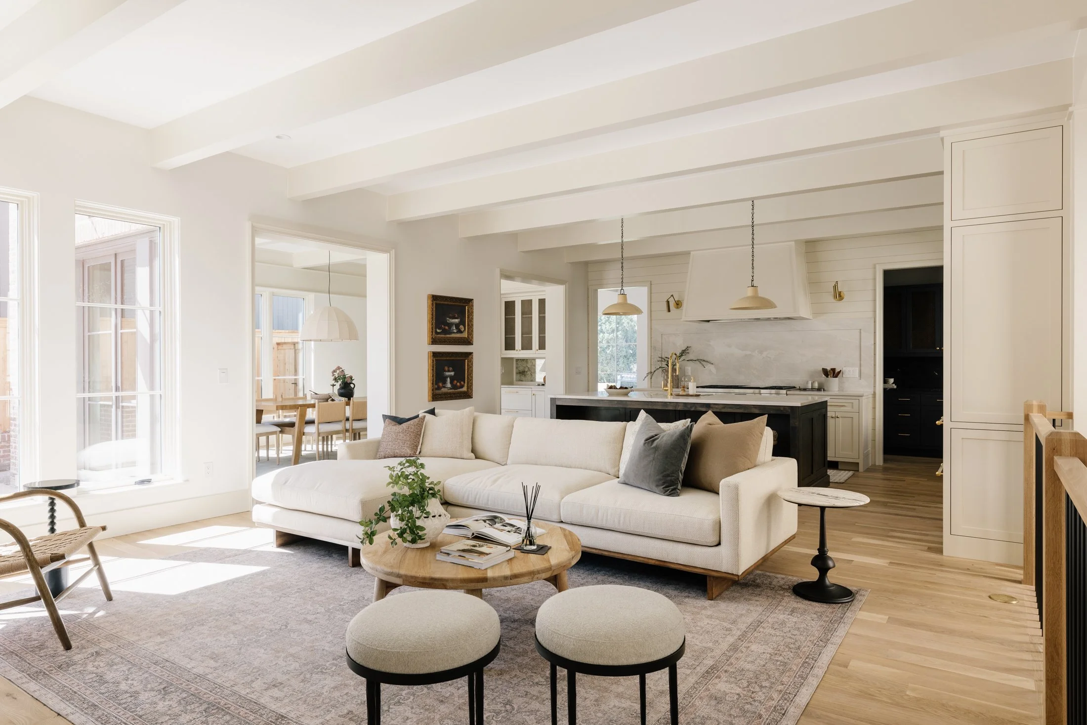 Bright living room with white sofa, round coffee table, and view into dining and kitchen areas with light wood floors and large windows.