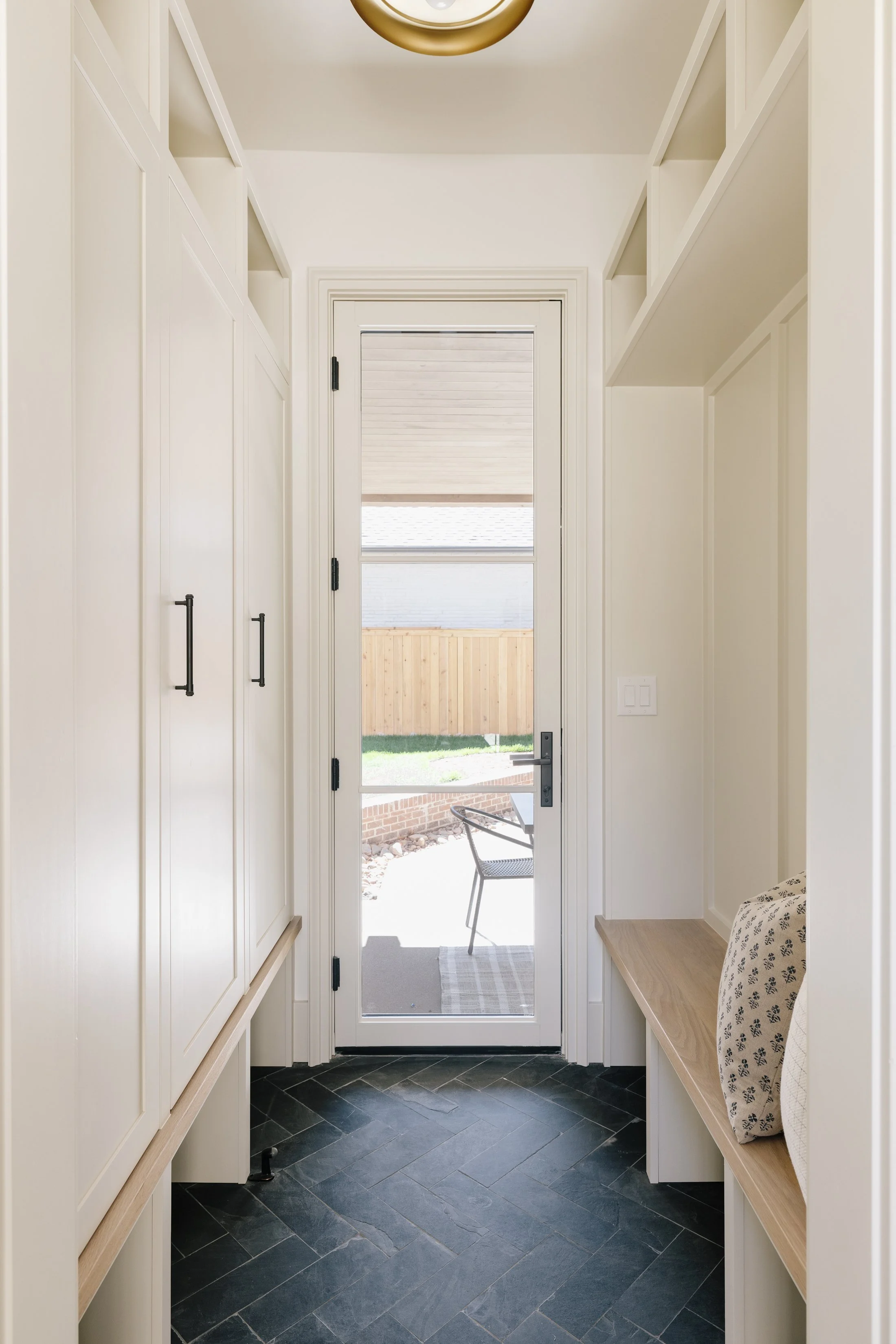 A small entryway with white cupboards on the left, a wooden bench on the right with a white pillow, black handles on the cupboards, a glass door leading outside, and a black slate tiled floor.