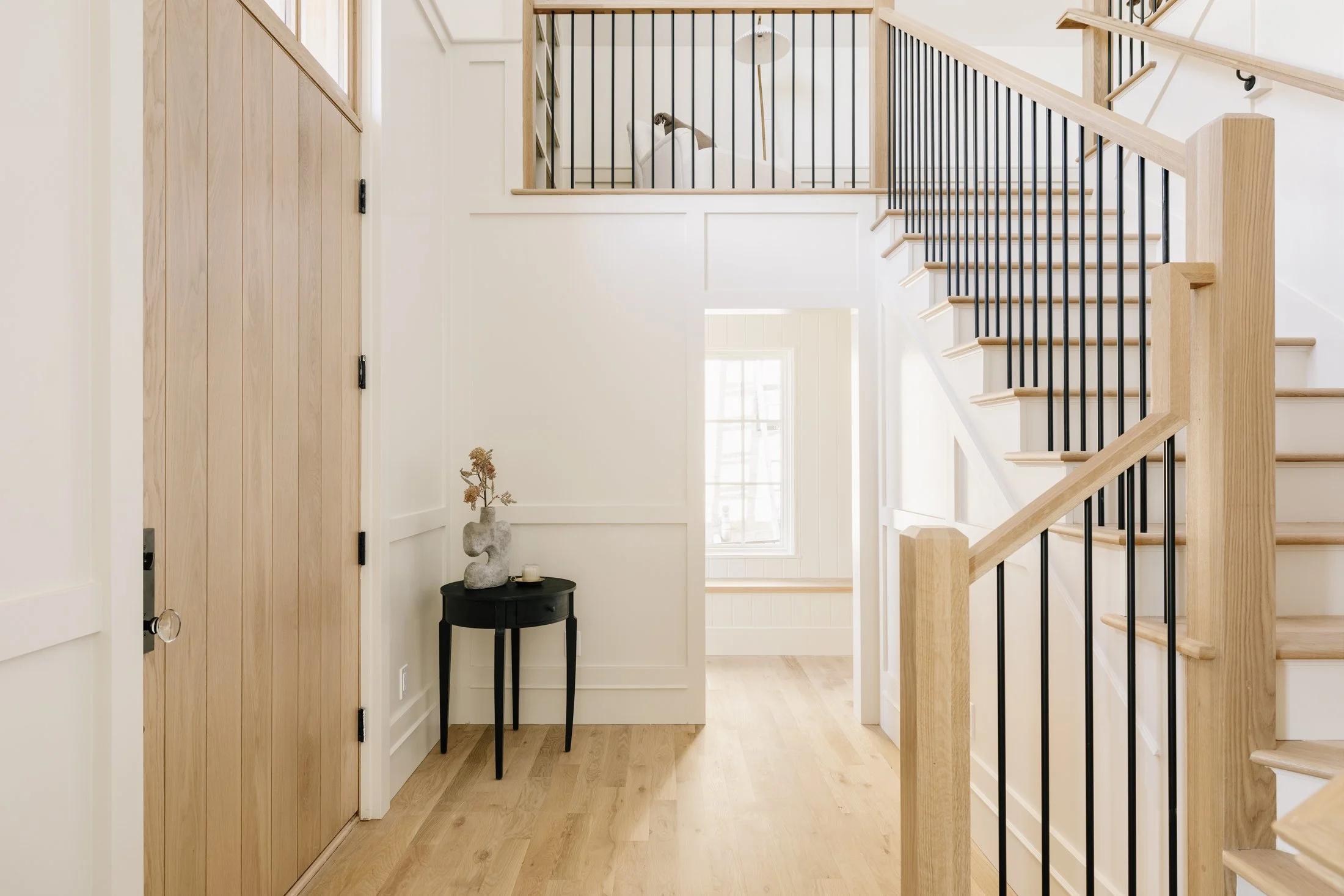 Interior of a modern home's stairway with wooden steps and black metal balusters, a small black side table with a vase and flower, and a window in the background.