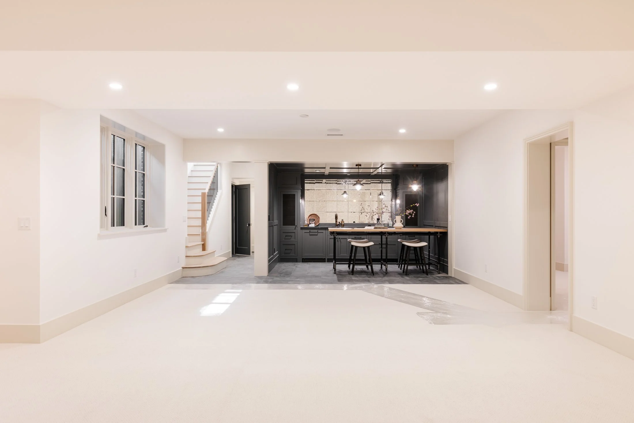 Empty living room with white walls and beige carpet, open kitchen with black cabinetry and barstools, staircase to the left, windows on the left wall, and ceiling lights.
