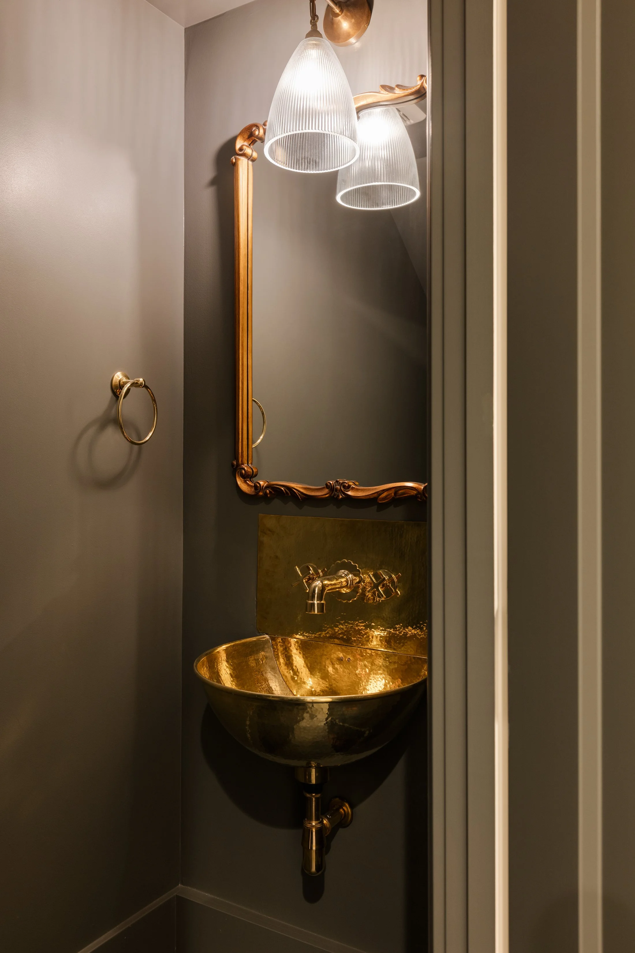 A small, gold metallic sink with two handles and a matching backsplash, mounted on a dark gray wall. Above it is an ornate gold-framed mirror and a modern pendant light fixture with two frosted glass shades. A towel ring is attached to the wall on th