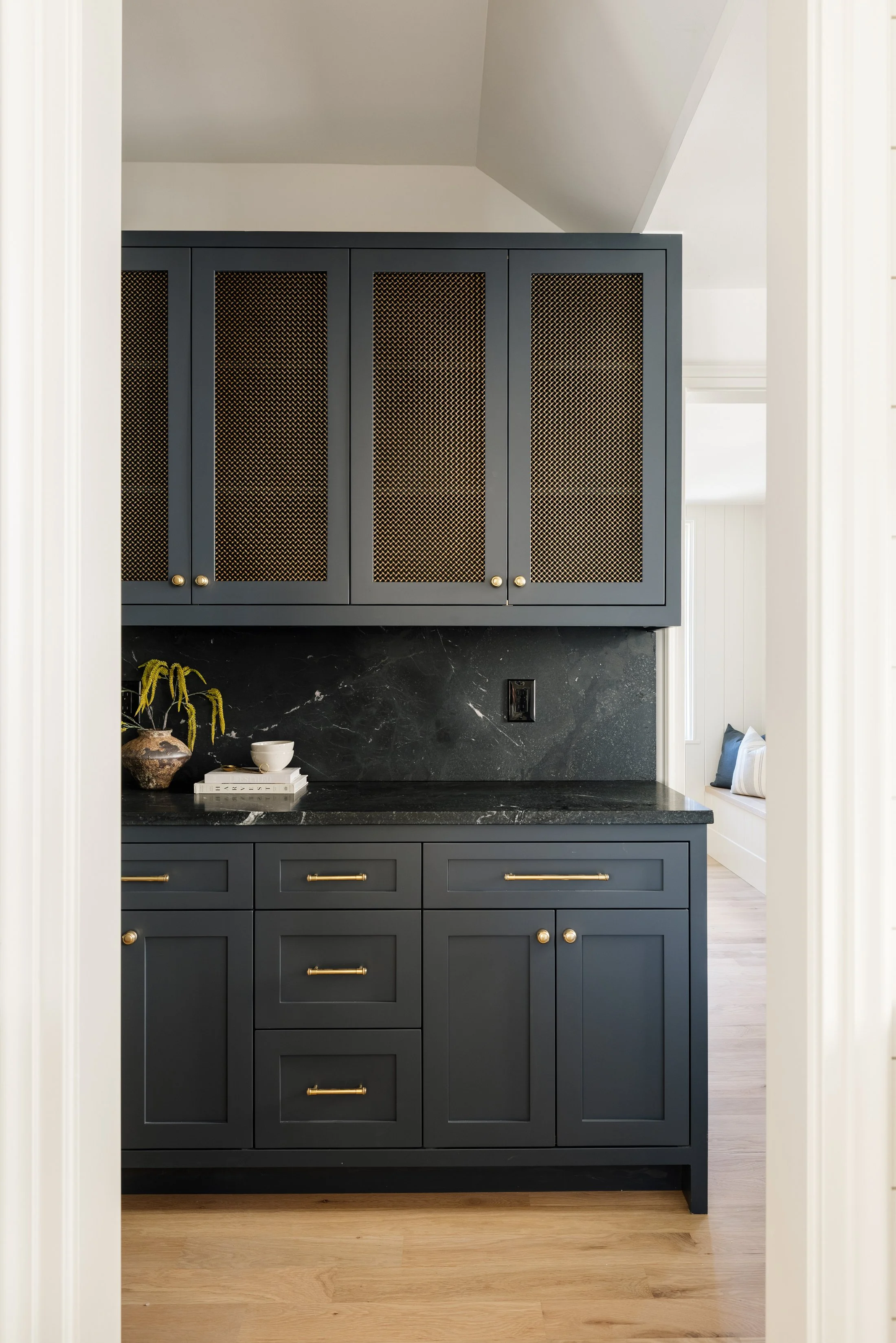 A kitchen cabinet painted in a dark blue color, with gold hardware, featuring upper cabinets with wire mesh doors and lower drawers and cabinets with solid doors. There is a black marble countertop with a decorative vase, a white bowl, and some books