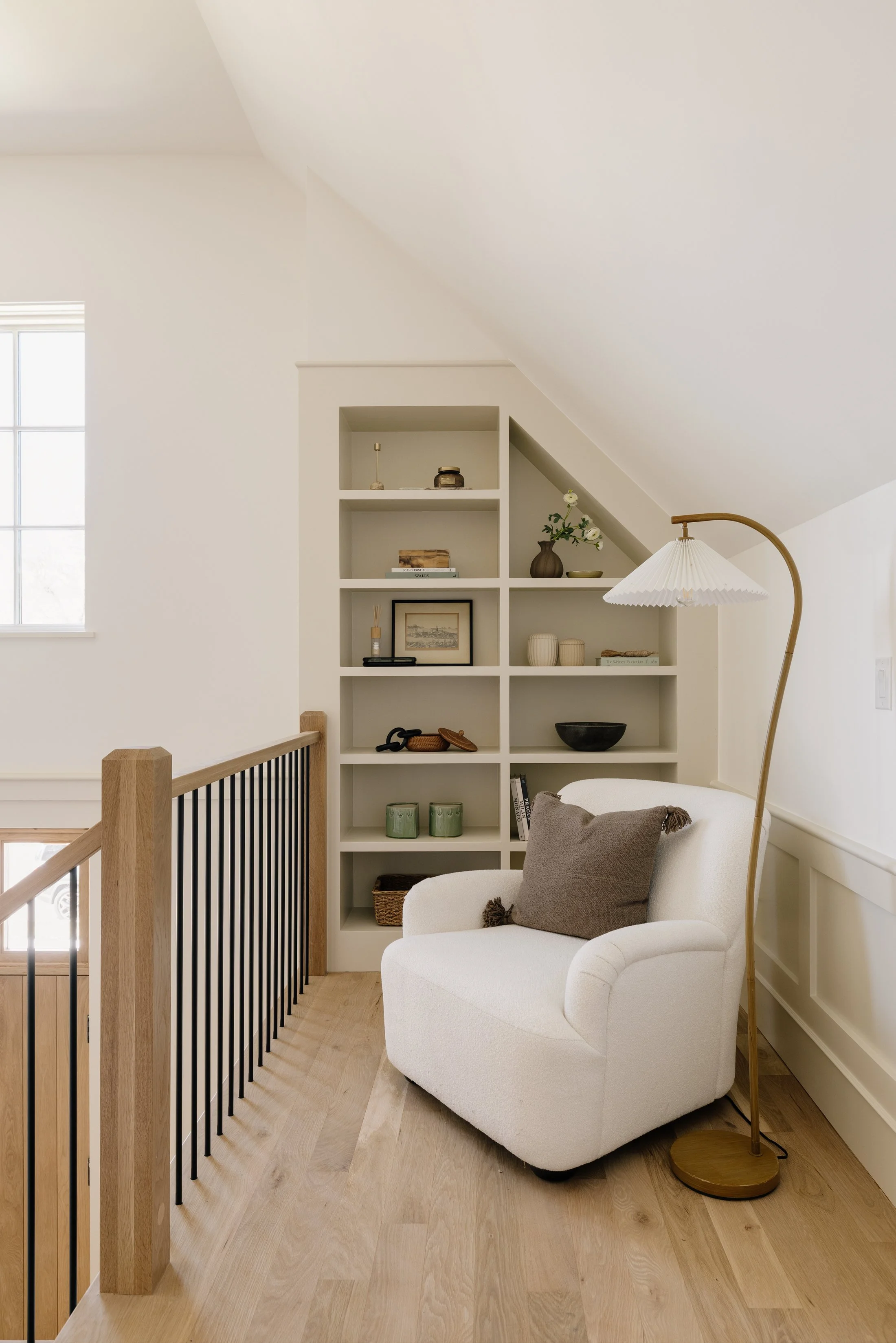 A cozy corner with a white armchair and a brown pillow next to a tall floor lamp with a white lampshade and brass base. Behind the chair is a built-in shelf filled with decorative items, framed pictures, and books. A window to the left provides natur