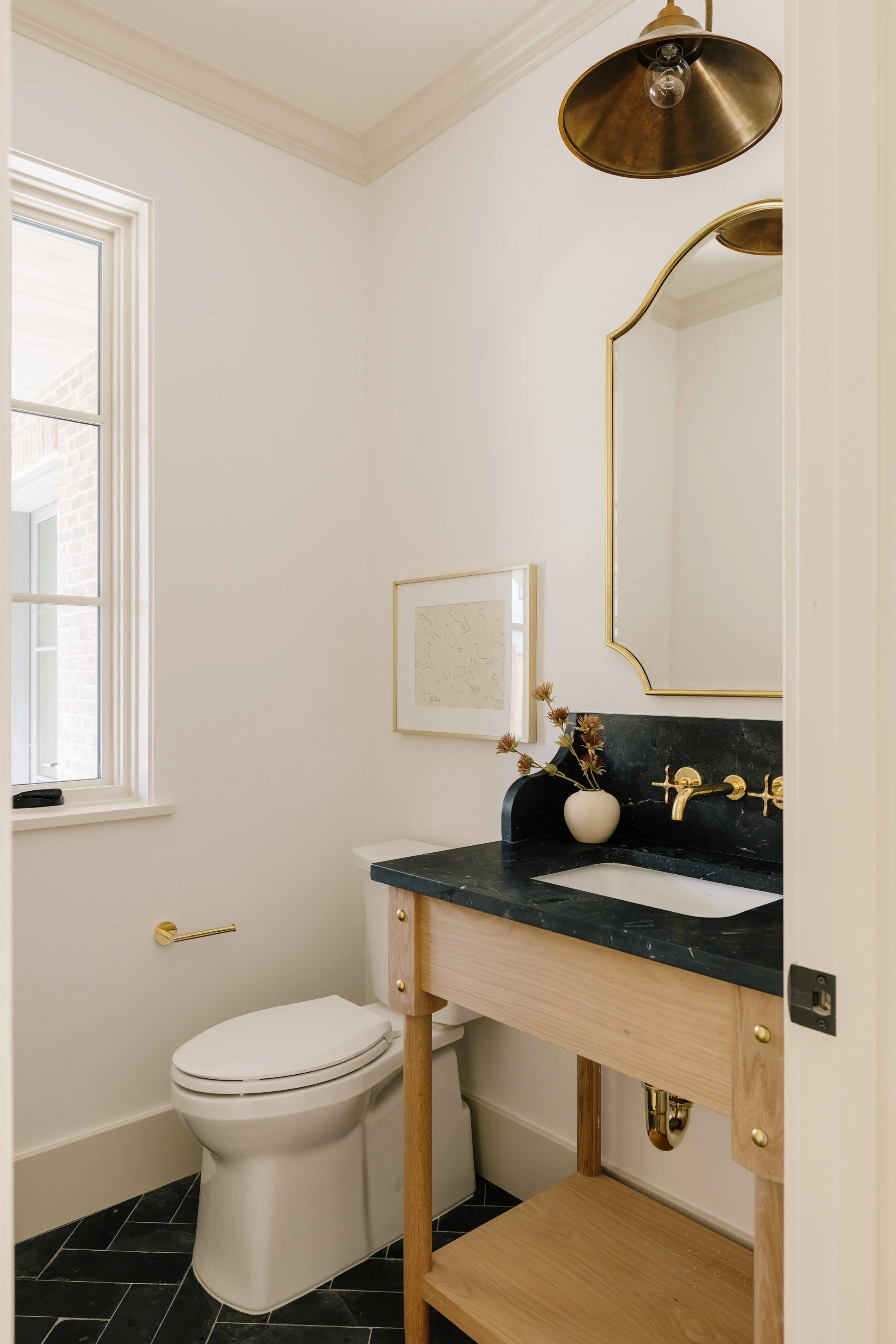 A small bathroom with a white toilet, a black marble vanity with a gold faucet, a minimalist framed art piece on the wall, and a window letting in natural light.