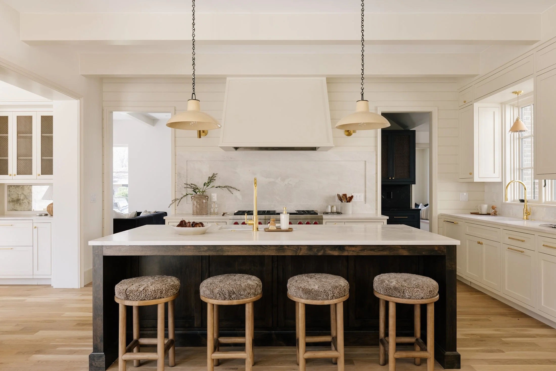 Modern kitchen with white cabinets, marble countertops, a kitchen island with a dark wood base, four beige upholstered stools, and gold accents on fixtures and handles.