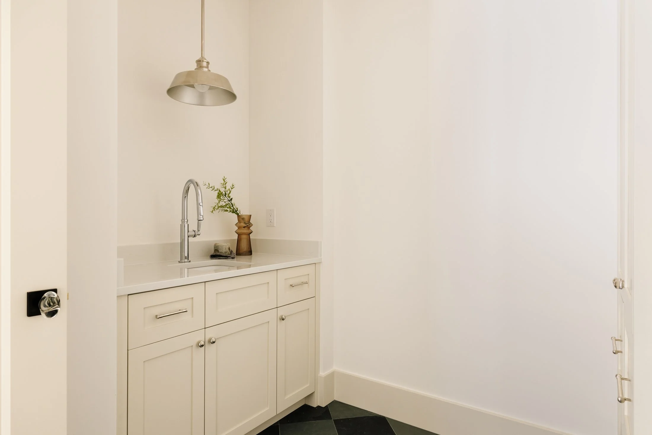 Small white kitchenette with a stainless steel sink, a faucet, a wooden vase with greenery, a black soap or sponge, a hanging pendant light, white cabinets, and a black and white tiled floor.