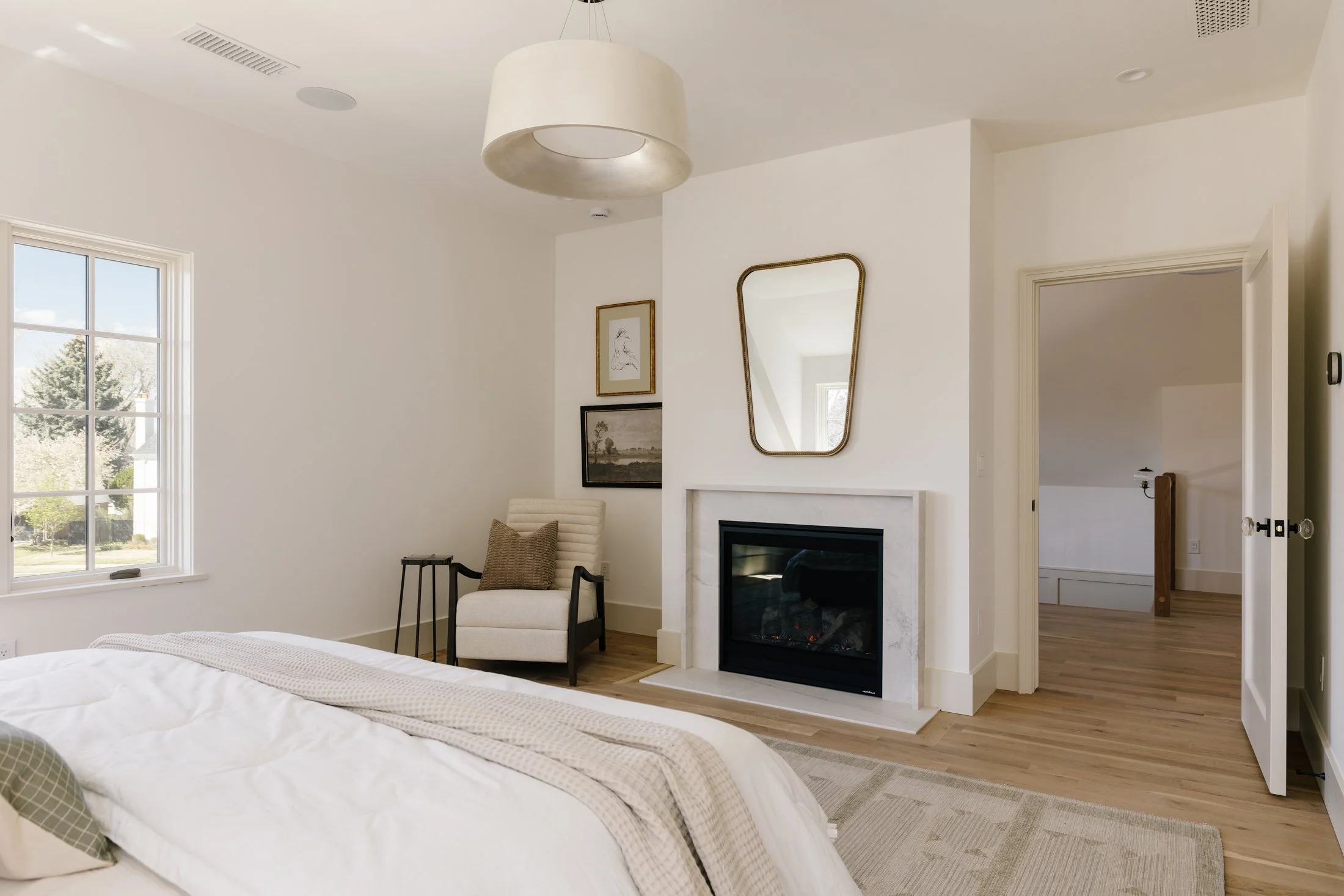 Bright bedroom with white walls, a large window, a heated fireplace with a mirror above, and a cozy chair in the corner.