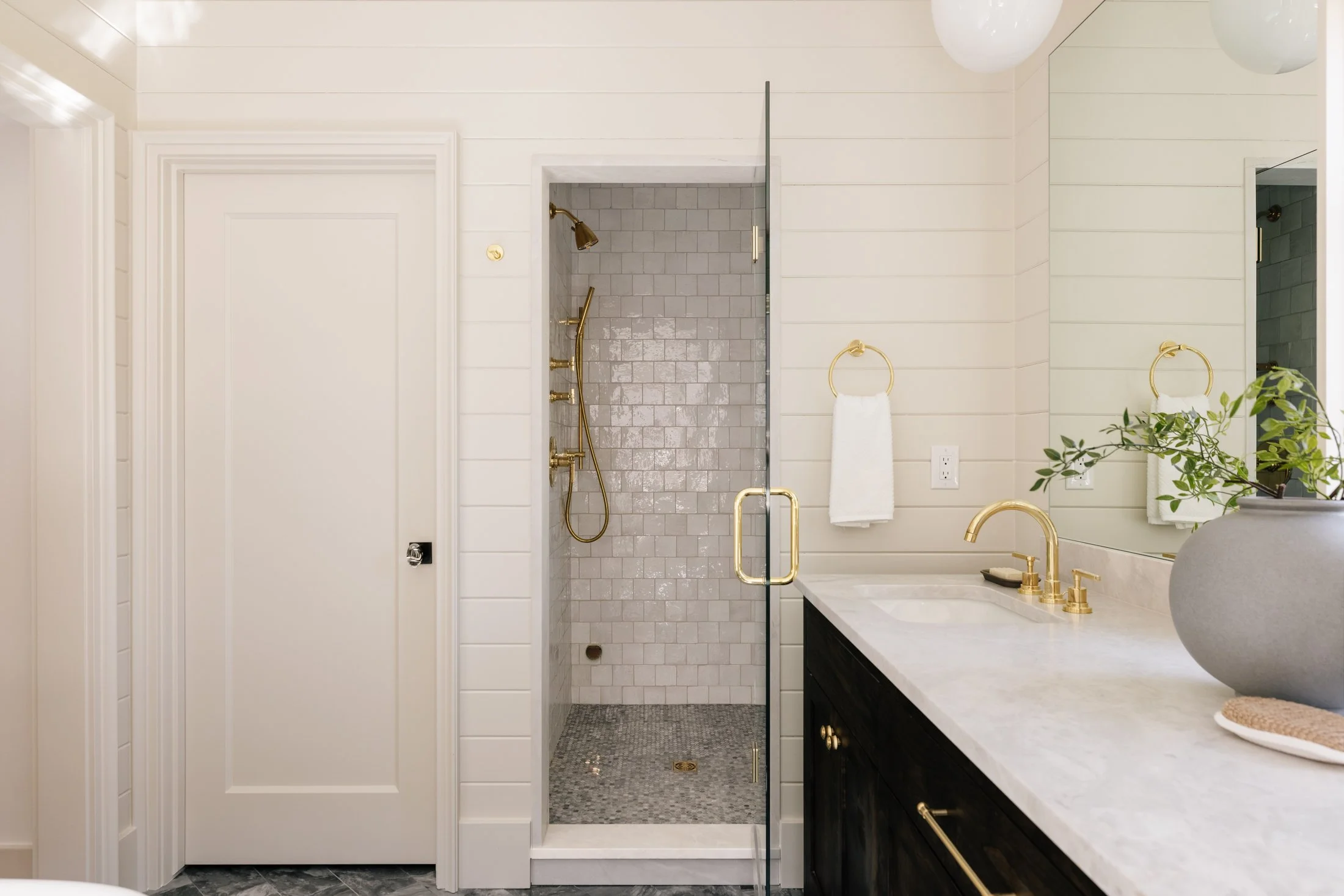 Modern bathroom with white paneled walls, a glass shower door, gold fixtures, a marble countertop with a sink, and decorative plant in a vase.