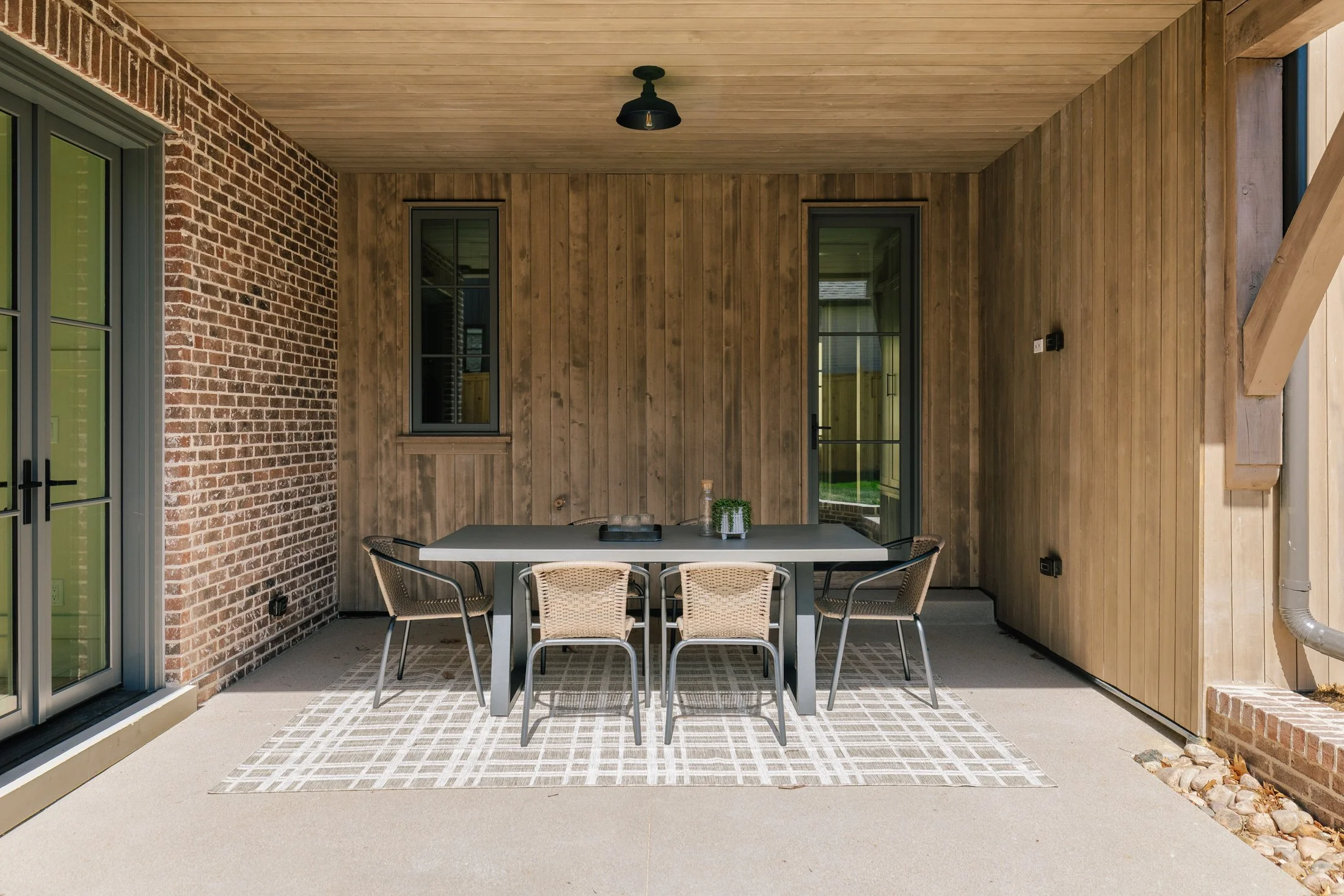 Outdoor patio with a black dining table and six woven chairs, brick and wood siding, two windows and a glass door, decorated with a small plant and candle holders, with a patterned outdoor rug, on a concrete floor.