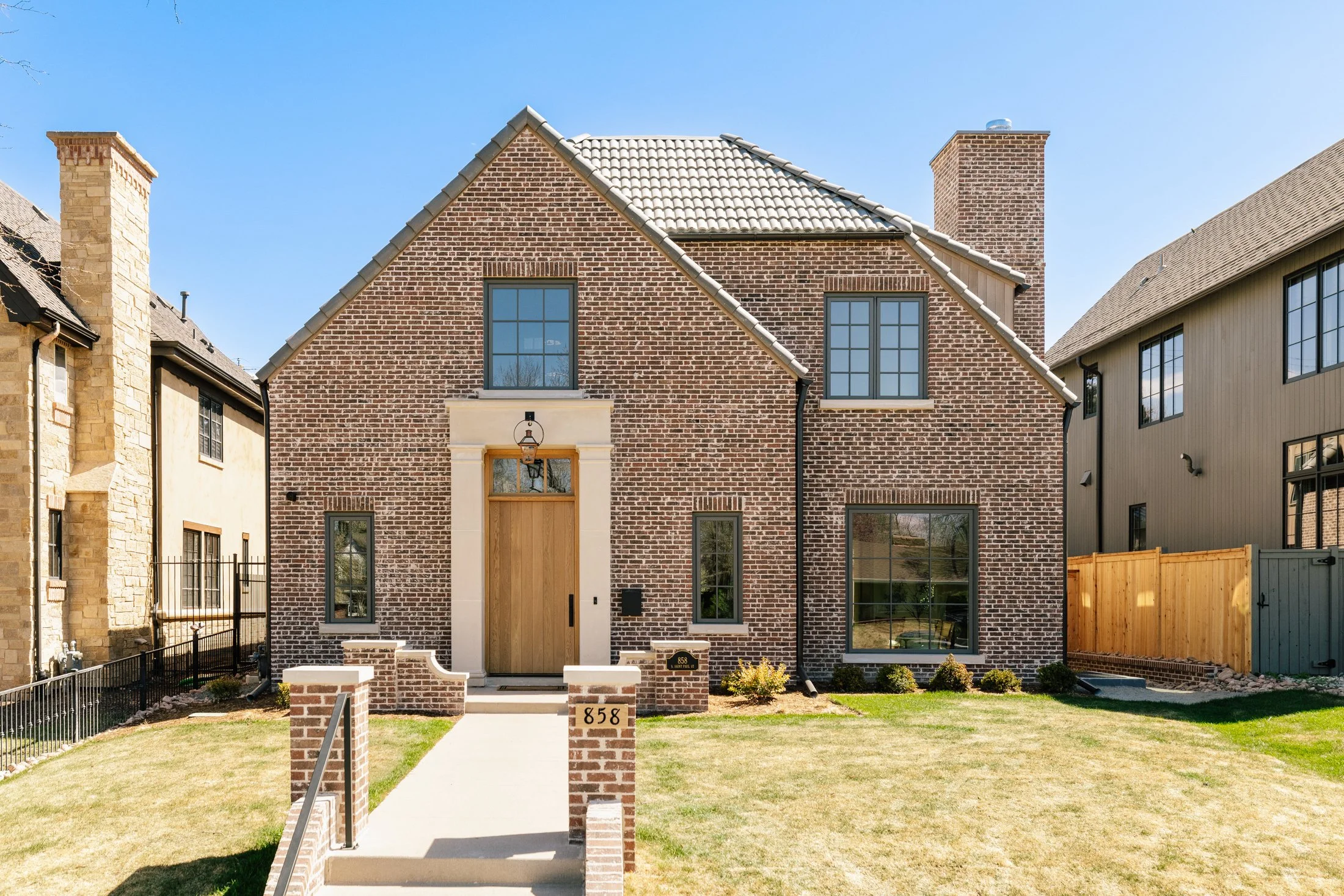 Front view of a modern brick house with a wooden front door and large windows, situated on a manicured lawn with a fence and neighboring houses on either side.