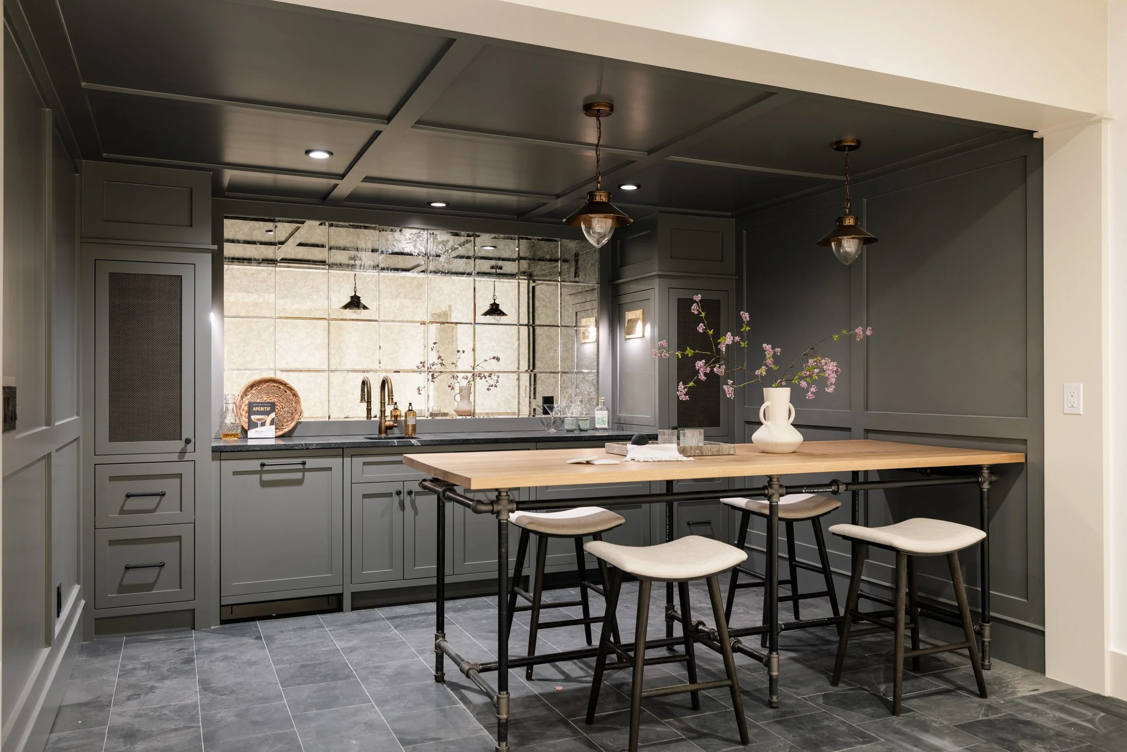 Kitchen with green cabinets, a wooden island with four stools, black countertops, a mirrored backsplash, and pink flowers in a white vase.