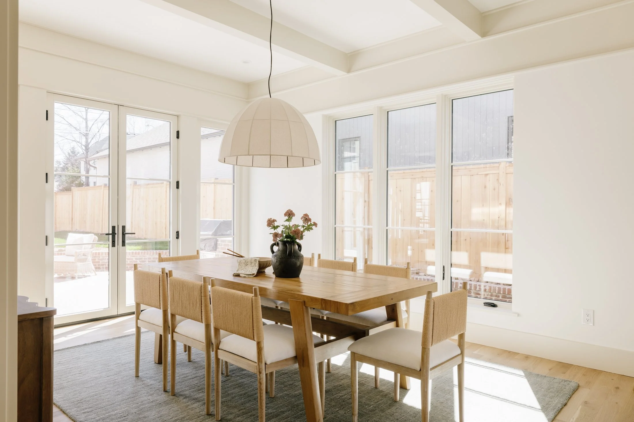 Bright dining room with a wooden table, eight beige chairs, large windows, and a vase with flowers on the table.