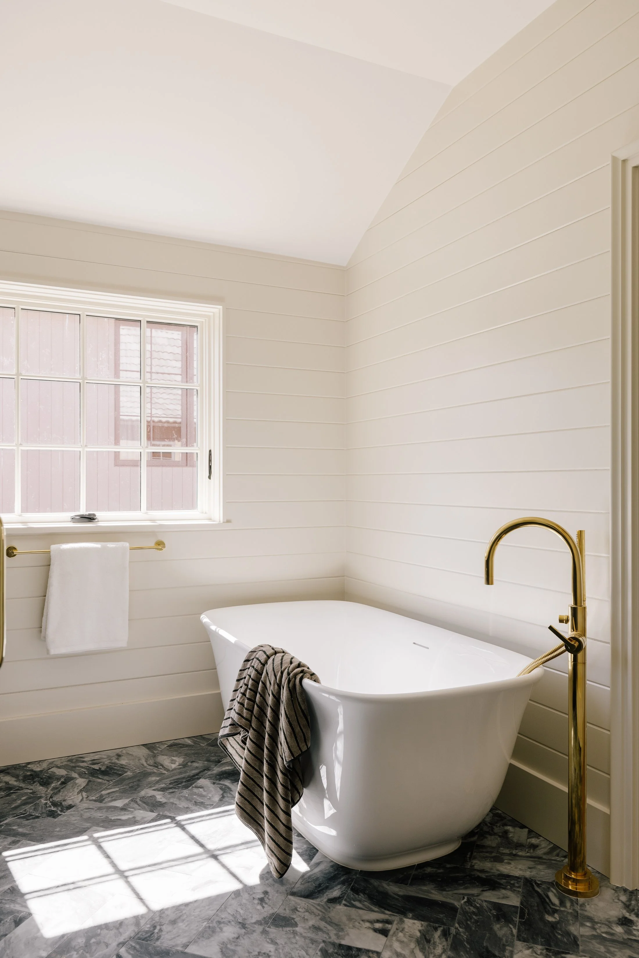 A white freestanding bathtub with a gold faucet in a bright bathroom with white shiplap walls, a window with multiple panes, a gold towel bar with a white towel, and a black and white striped towel hanging over the tub, with marble flooring.