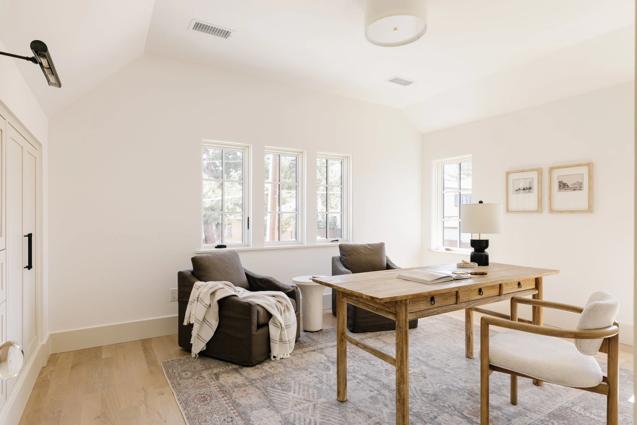 Bright living room with white walls, hardwood floor, a wooden table, two brown armchairs with blankets, a cream-colored armchair, a lamp, and framed pictures on the wall.