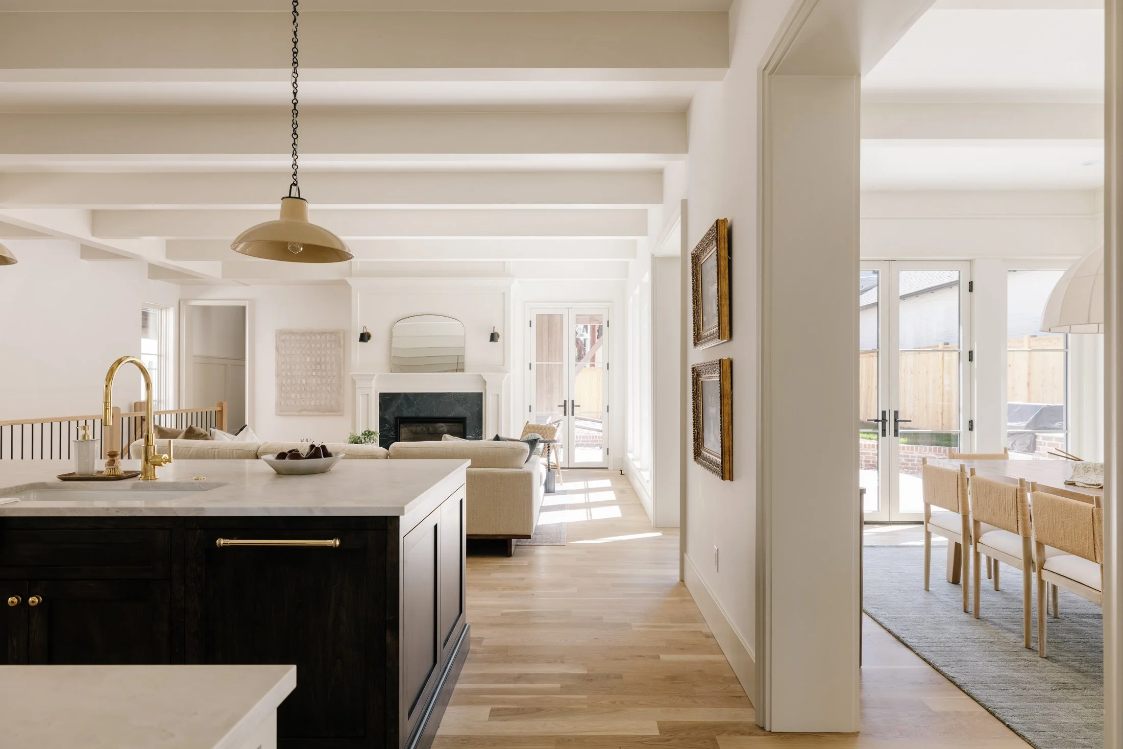 Open-concept kitchen and living area with white walls, wooden floors, and large windows letting in natural light. The kitchen island has a black base with a white countertop and gold fixtures. The living room has a beige sofa and a fireplace with a m
