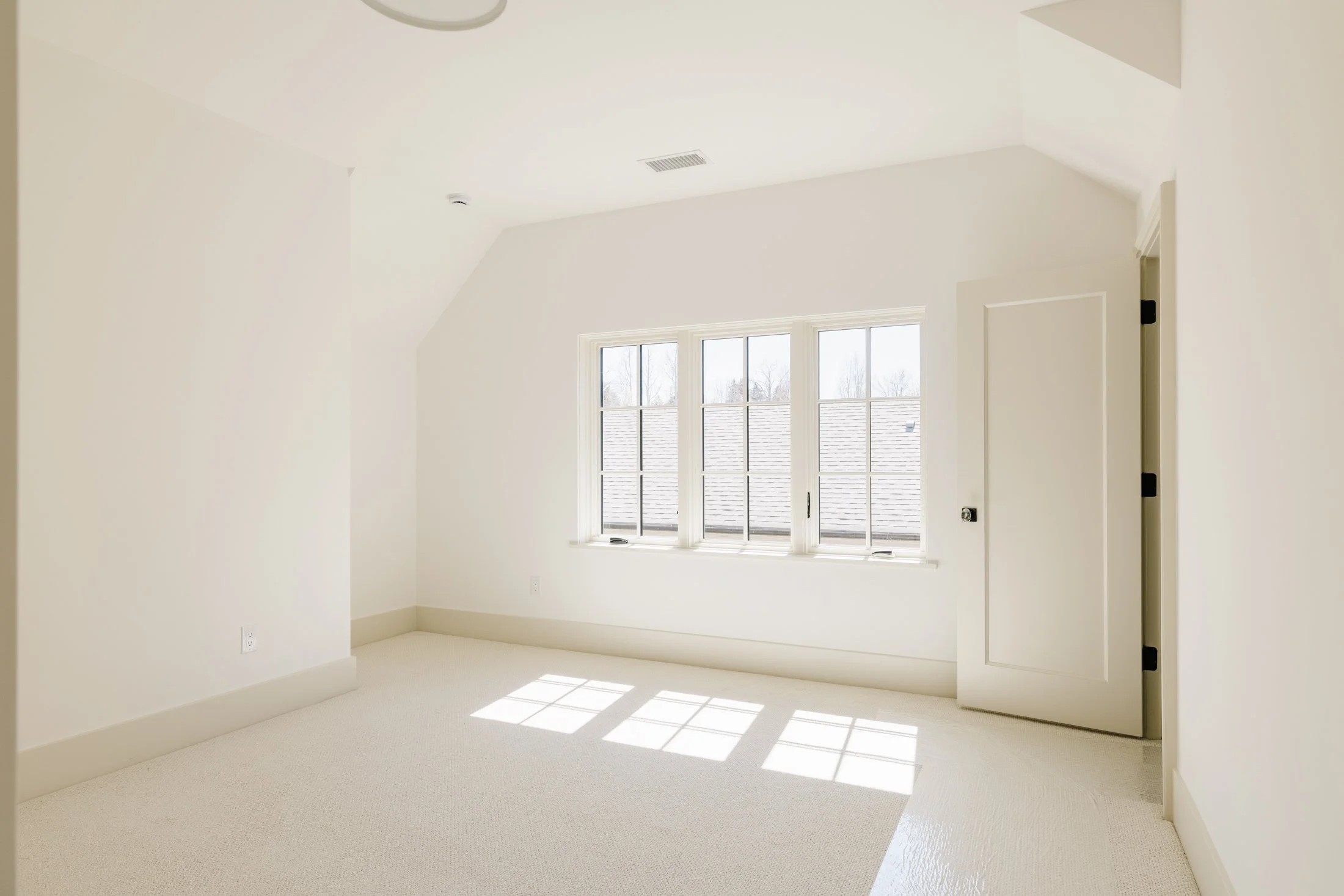 Empty room with white walls, carpeted floor, a large window, and a closed door.