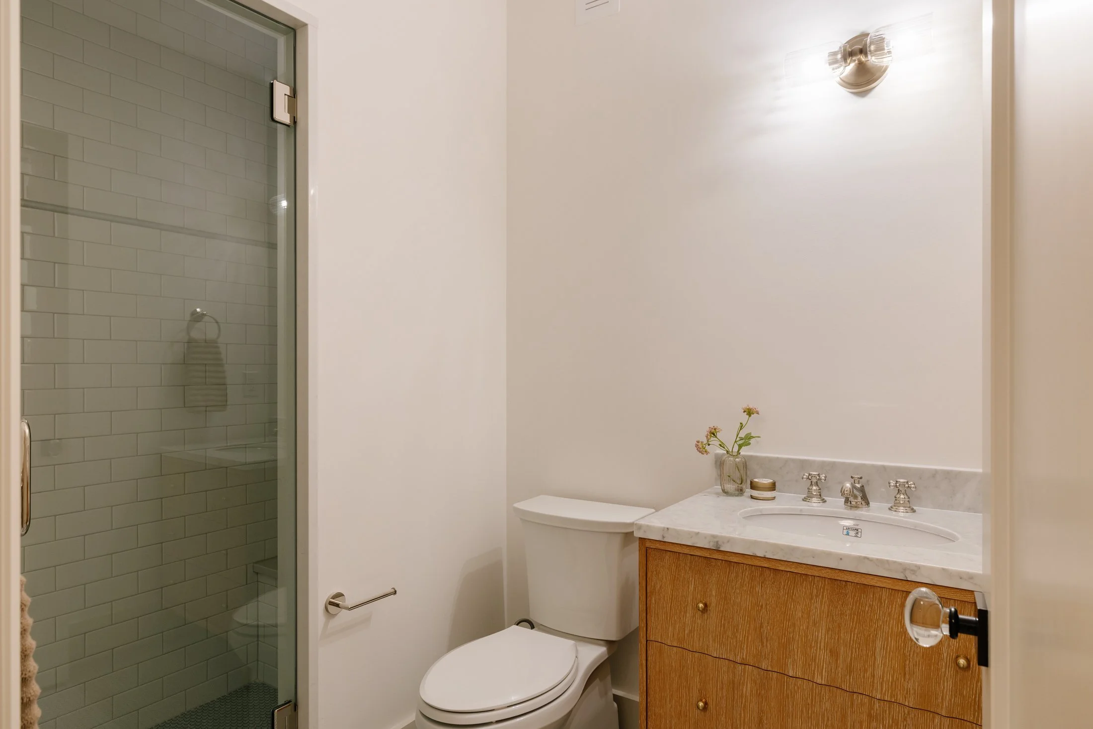 A small bathroom with a glass shower door, a white toilet, and a wooden cabinet with a marble countertop and sink. There is a small flower vase, a soap container, and a faucet on the countertop, and a wall-mounted light fixture above.