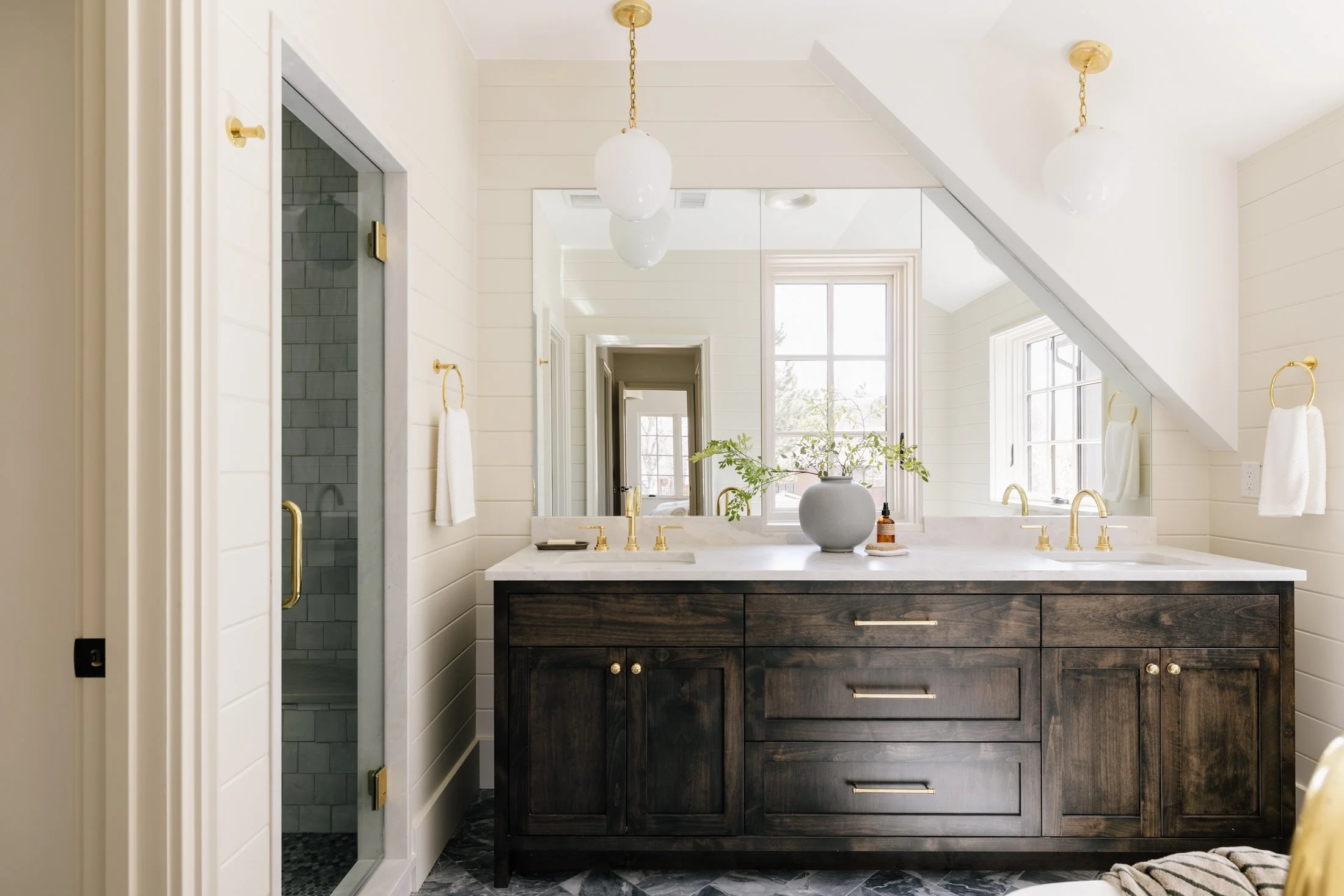 Bathroom with double sink vanity, dark wood cabinets, marble countertop, gold fixtures, large mirror, window, and a separate walk-in shower with glass door.