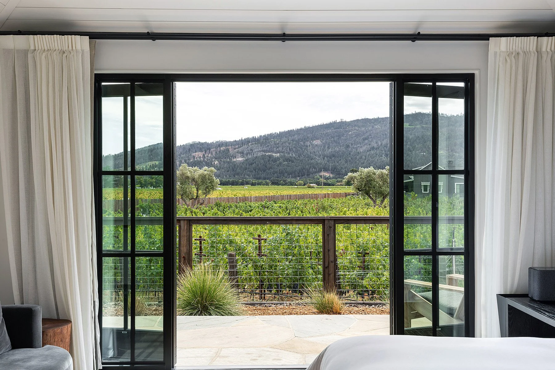 View through open sliding glass doors to vineyard and mountains beyond on a patio with plants and wooden railing, seen from inside a bedroom with white curtains.