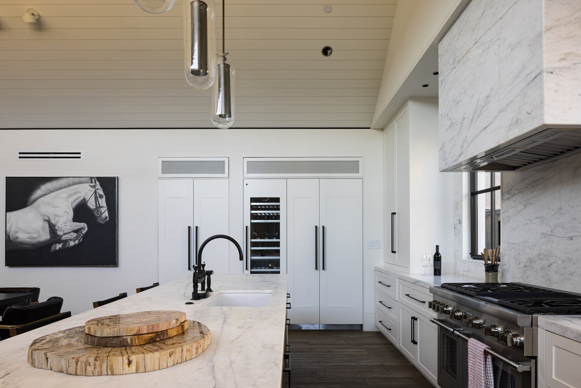 Modern white kitchen with marble countertops, black hardware, a black stove, and a wine cooler, decorated with a black and white horse painting and two wooden cutting boards on the island.