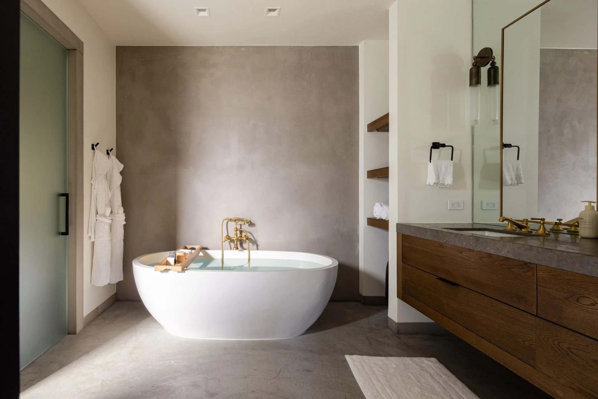 Modern bathroom with a freestanding bathtub, gold fixtures, a wooden vanity with a large mirror, and open shelving with towels.