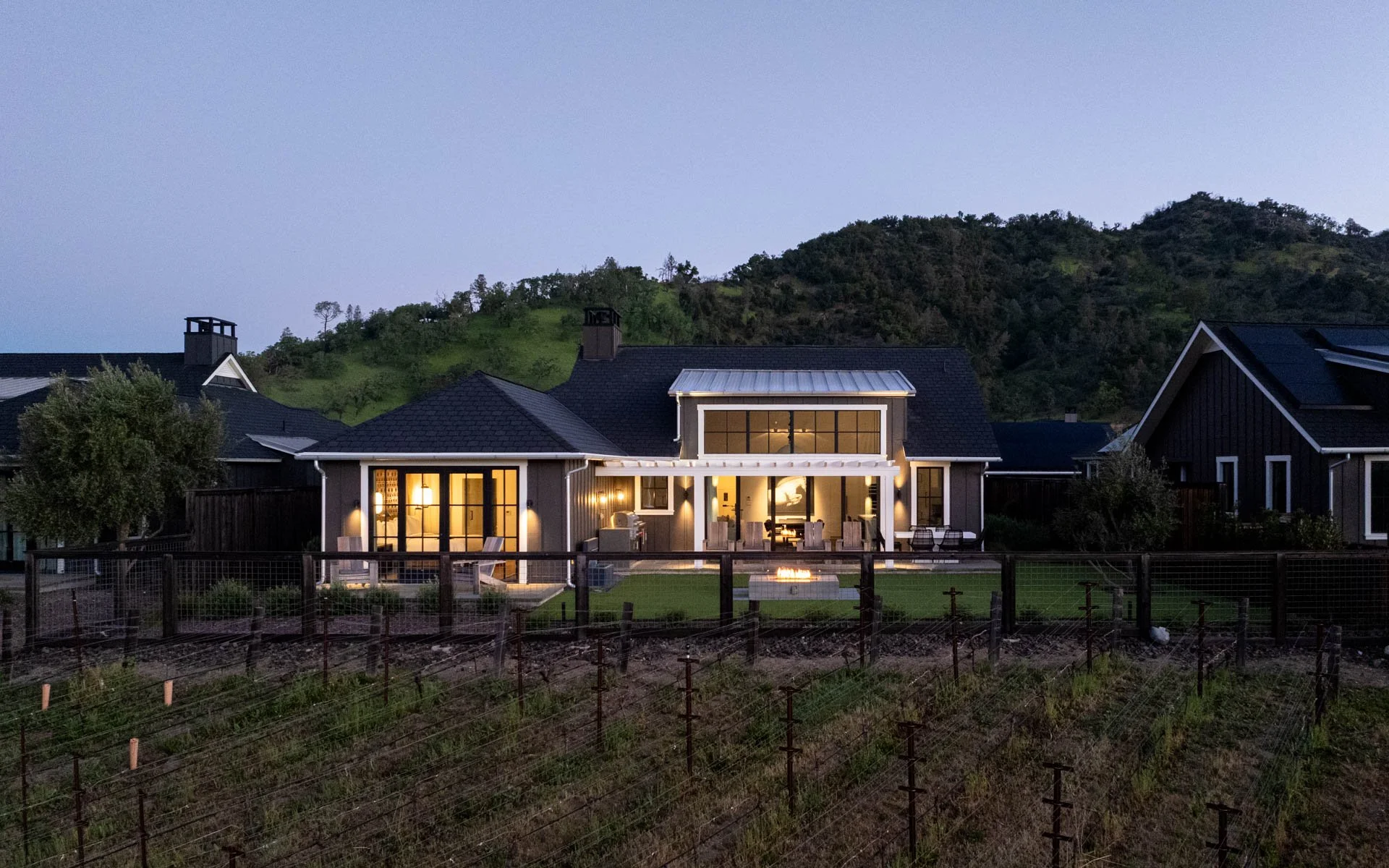 A modern house at dusk with large illuminated windows, outdoor seating, and a backyard with a small fire pit, set against a backdrop of green hills.