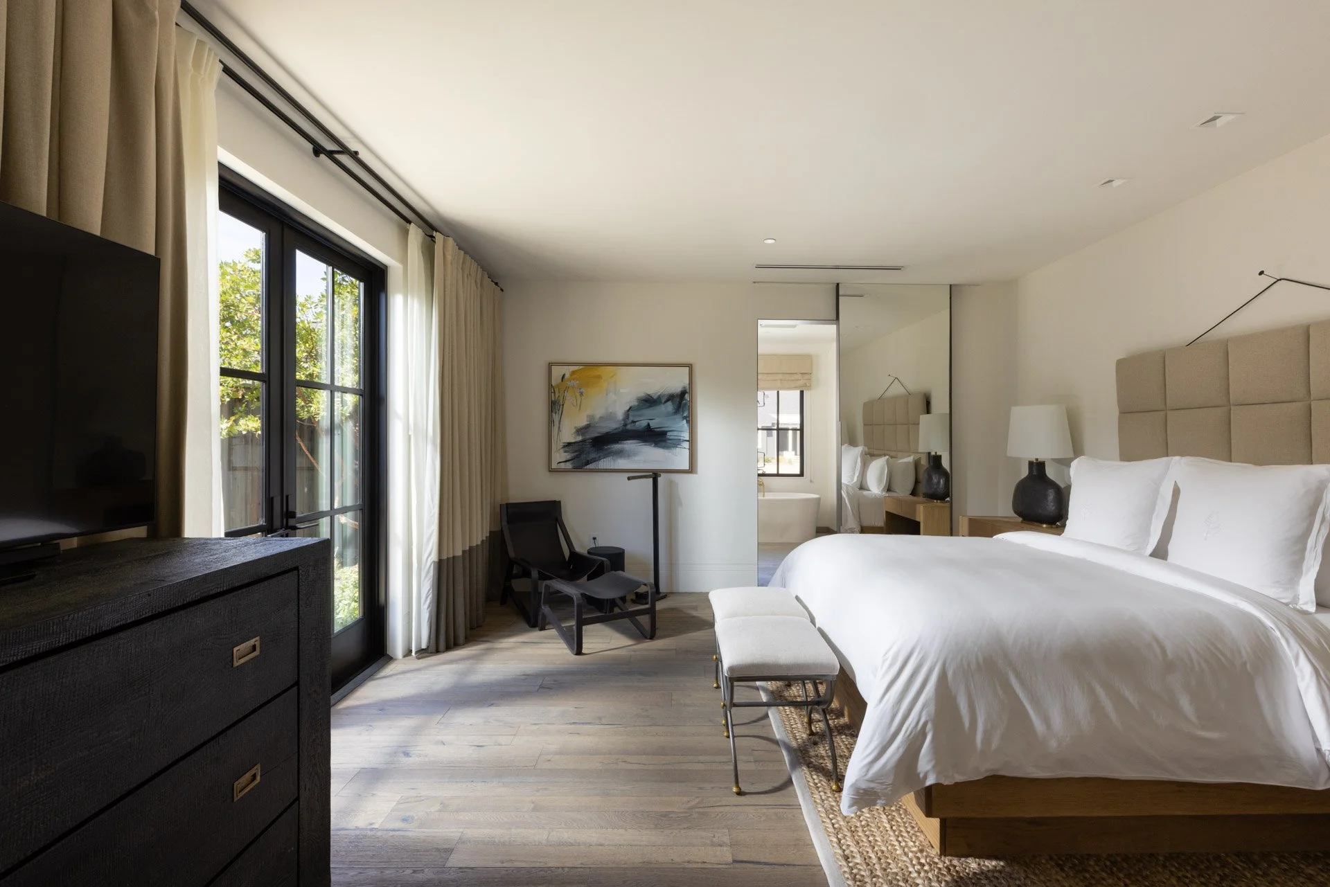 A modern hotel room with a large bed, white bedding, beige headboard, two black bedside lamps, a bench at the foot of the bed, a black chair, a colorful abstract artwork, a wide black TV stand, and sliding glass doors with beige curtains leading to a