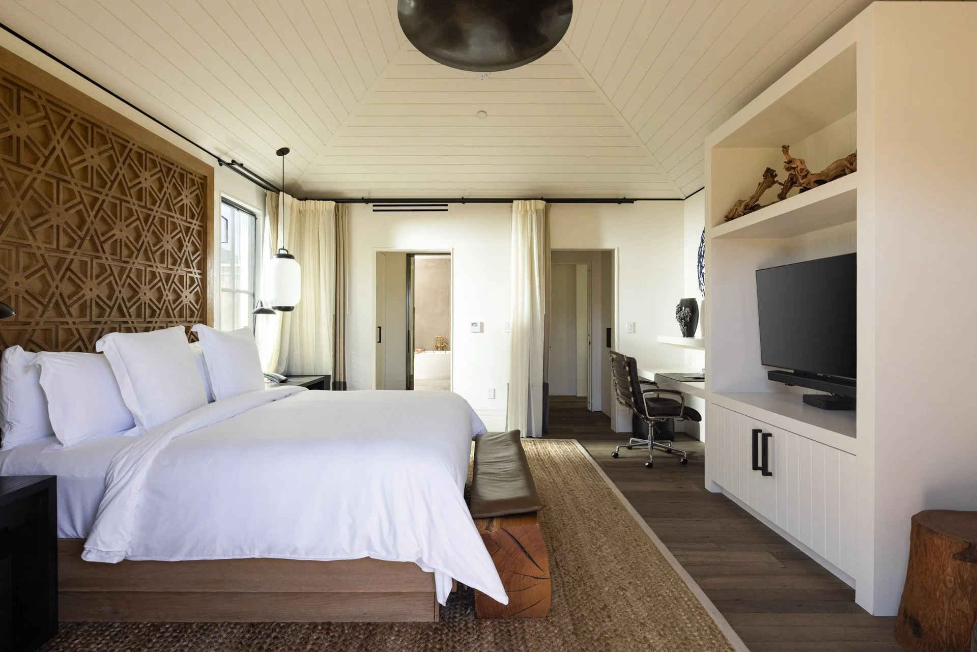 Modern bedroom featuring a large bed with white bedding, bedside lamps, a desk with a chair, a white storage unit with a TV, and decor accents, with neutral tones and wooden elements.
