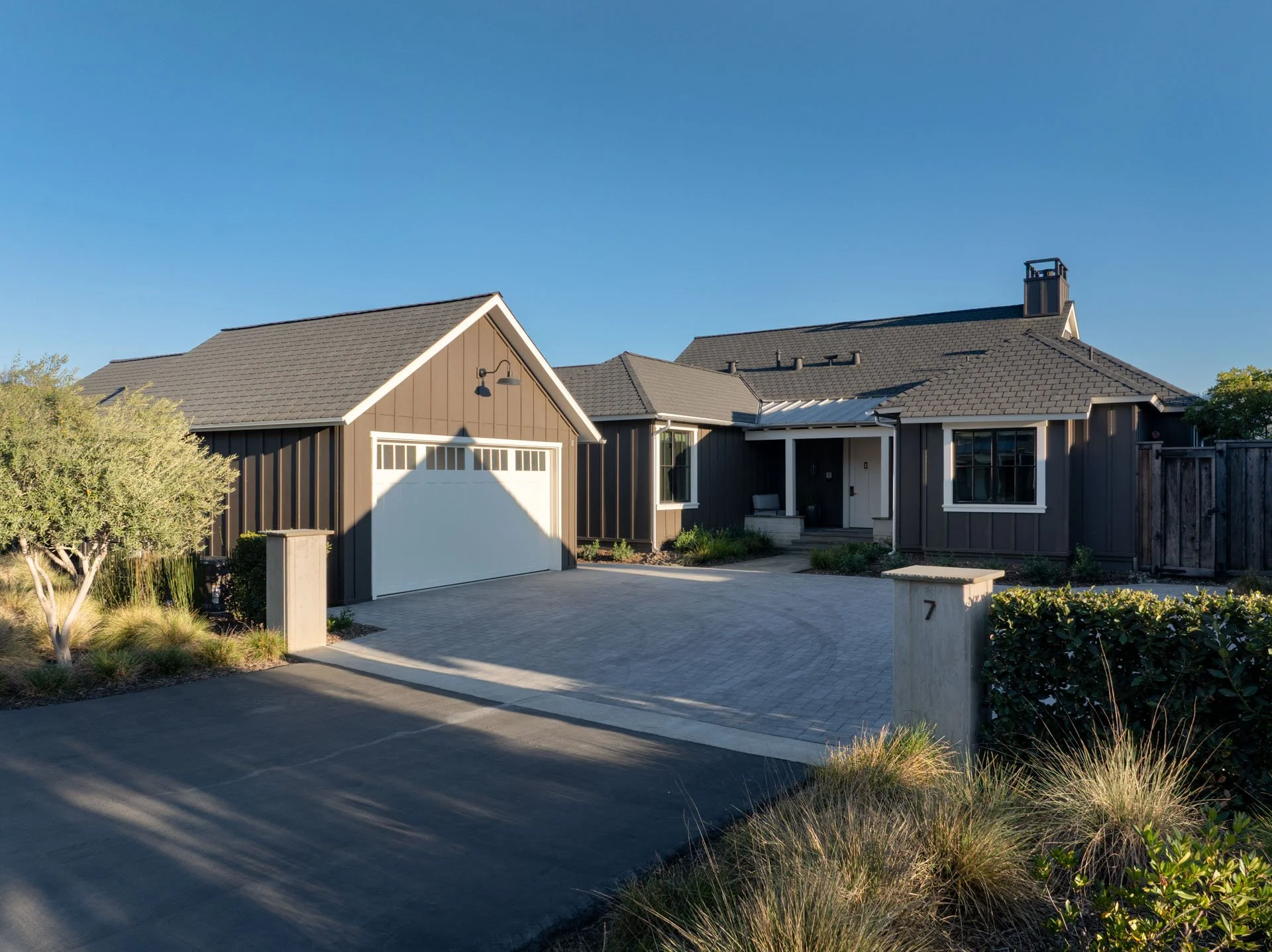 Front view of a modern house with a gray exterior, a white garage door, and landscaped yard with shrubs and plants under a clear blue sky.