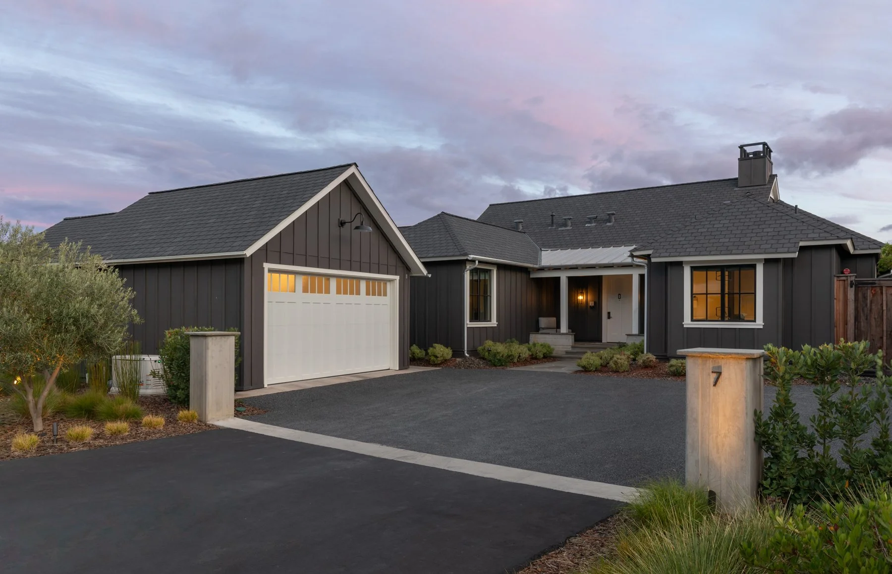 A modern black house with a garage, front porch, landscaped yard, and a driveway at dusk.