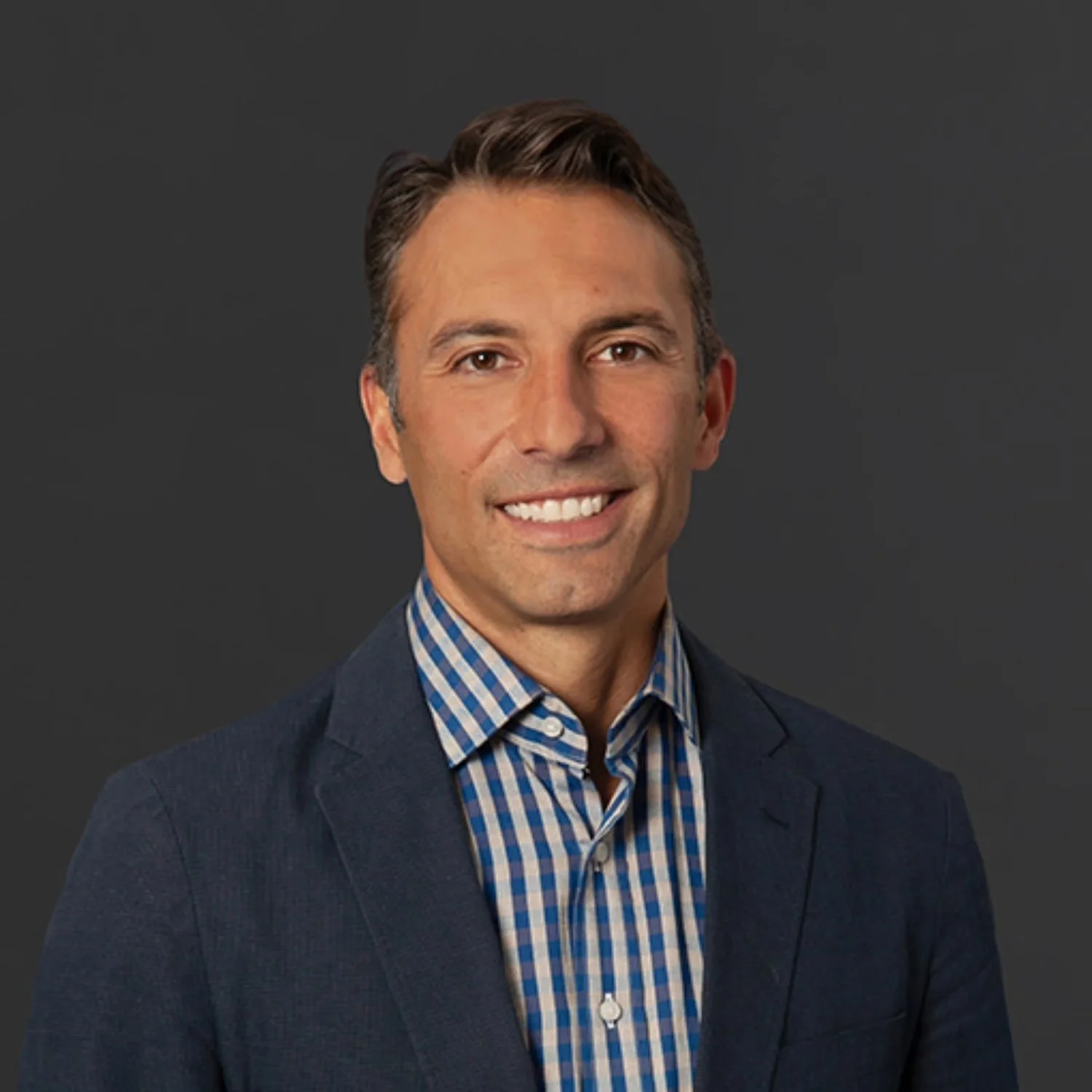 A man with short, dark blonde hair, wearing a navy blazer and a blue checkered shirt, smiling against a dark gray background.