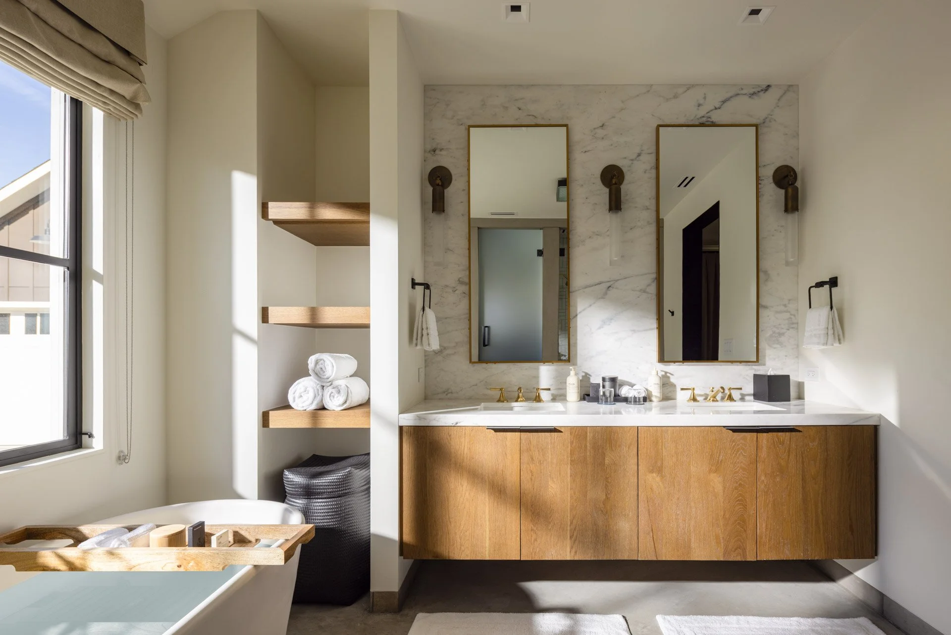 Modern bathroom with double vanity, marble backsplash, wooden cabinetry, two mirrors, and hanging sconces, next to a bathtub with rolled towels and a small wooden tray.
