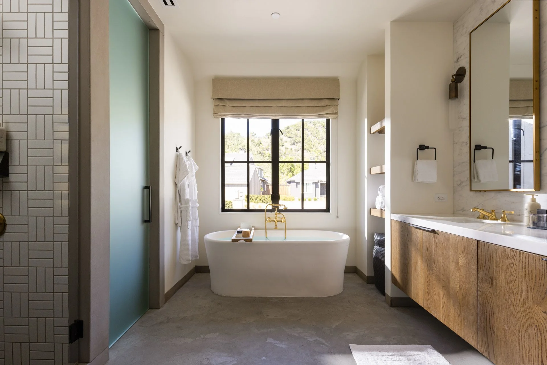 Modern bathroom with a bathtub in front of a large window, bathroom shelves with towels, a vanity with a mirror and gold fixtures, and minimal decor.