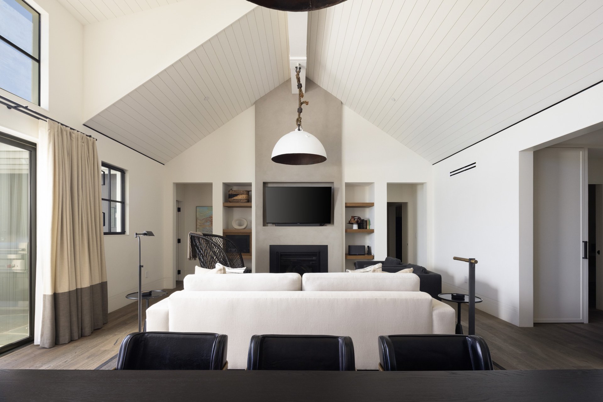 Living room with vaulted white ceiling, black-framed windows with beige curtains, a white sofa, a black lounge chair, a TV mounted above a fireplace, and open shelving with decor and books.