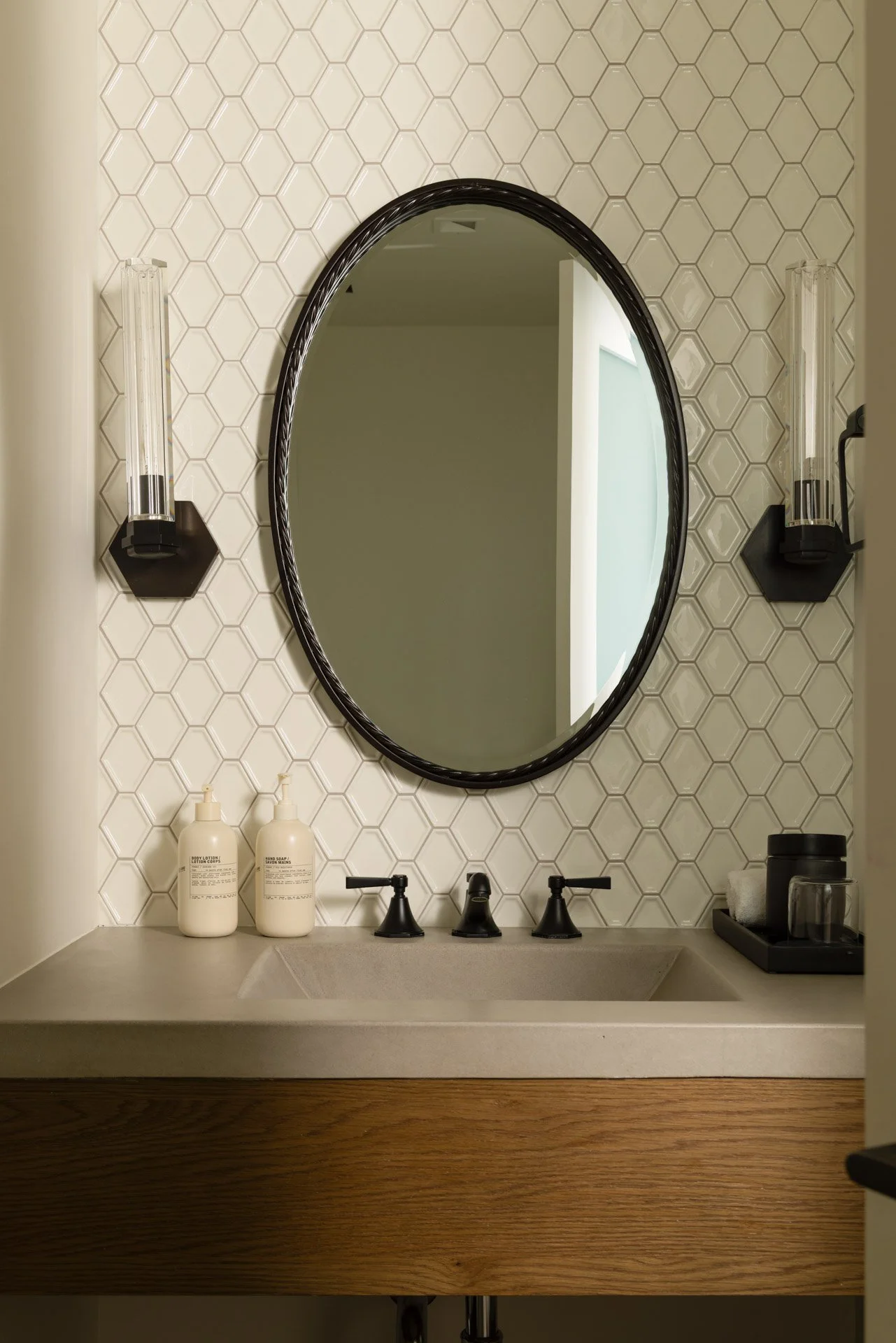A modern bathroom vanity area featuring an oval framed mirror, hexagonal white tile backsplash, black fixtures, and two bottles of soap or lotion on a concrete countertop. Two black wall-mounted light fixtures are on either side of the mirror, and a 