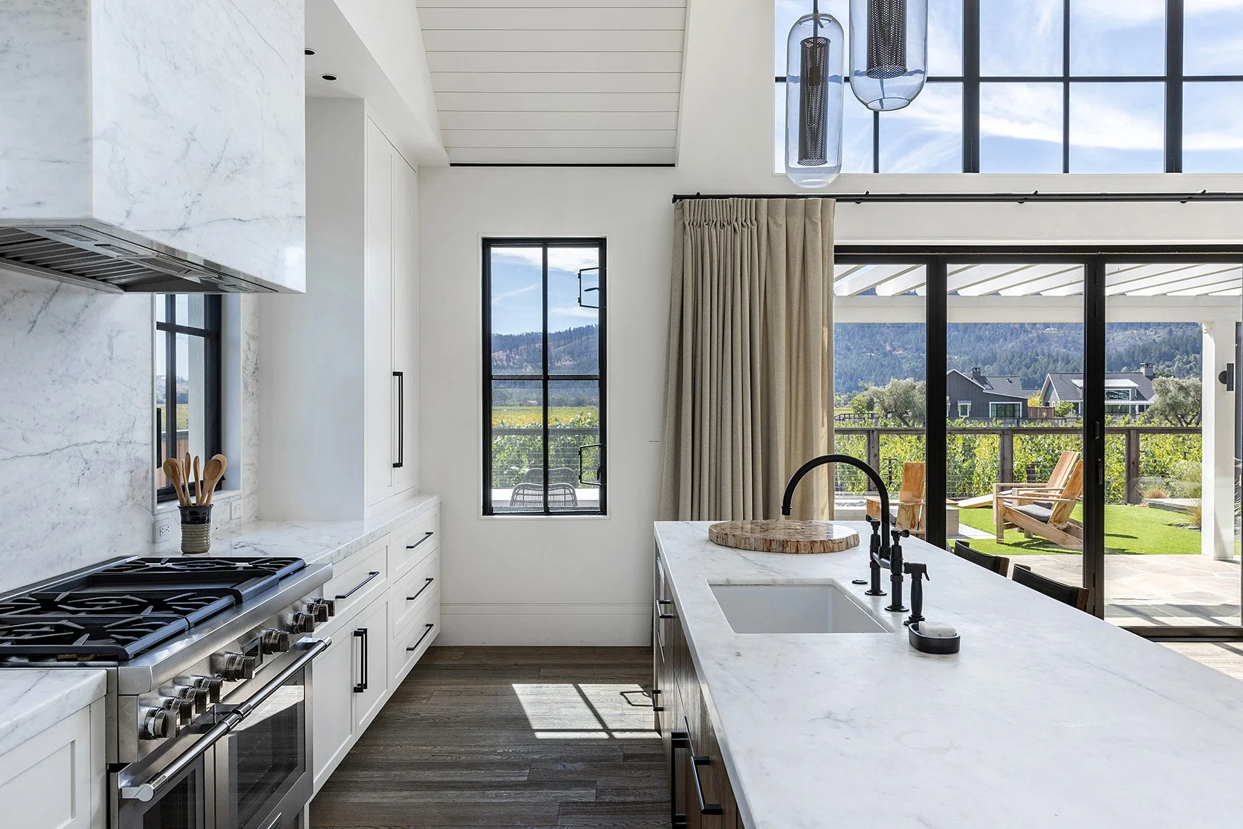 Modern kitchen with white marble countertops, a black faucet, and large windows overlooking a patio and scenic landscape.