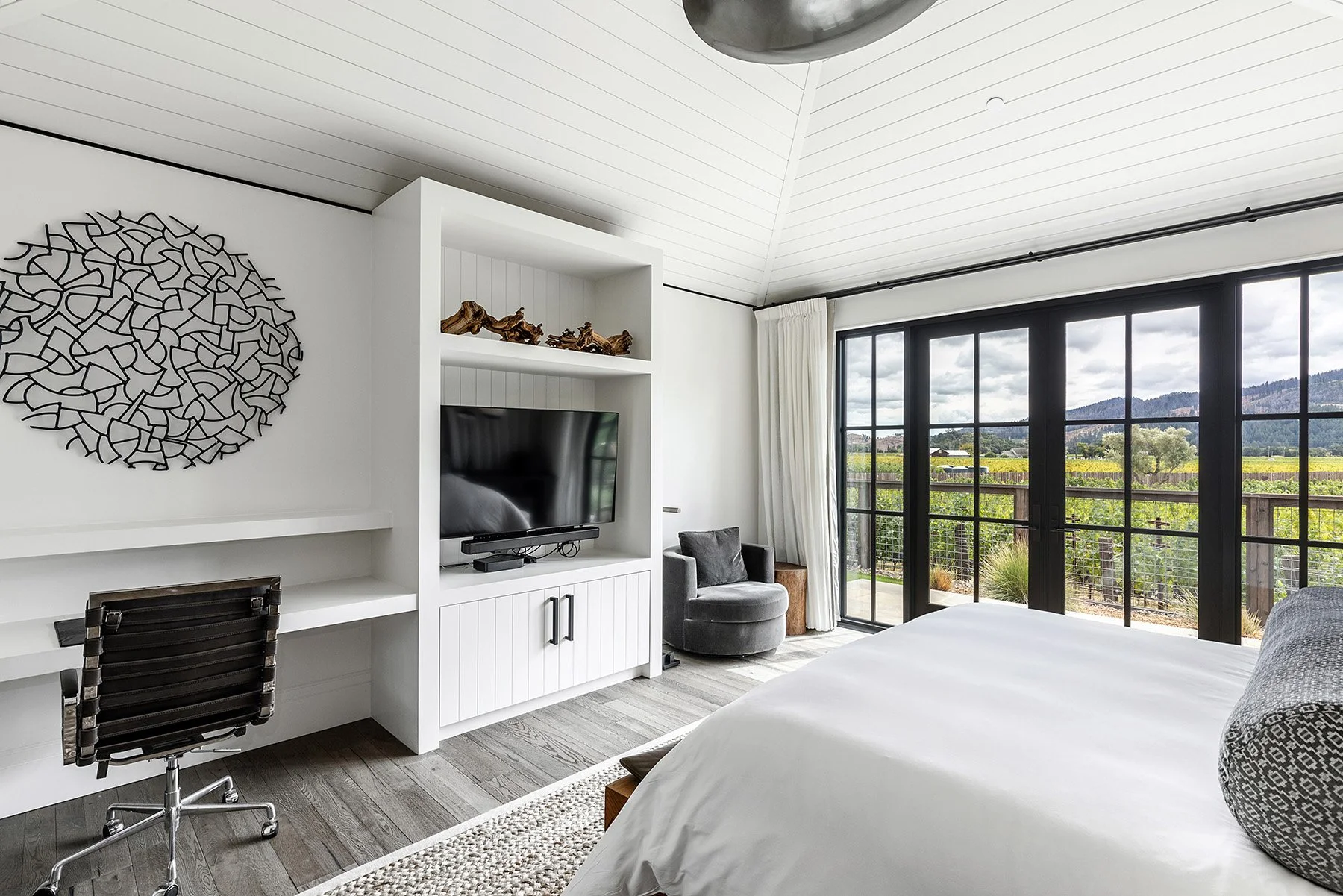 Modern bedroom with a large window overlooking a scenic outdoor landscape, a bed with a patterned pillow, a chair, a TV on a white cabinet, and decorative wooden sculptures on a shelf.