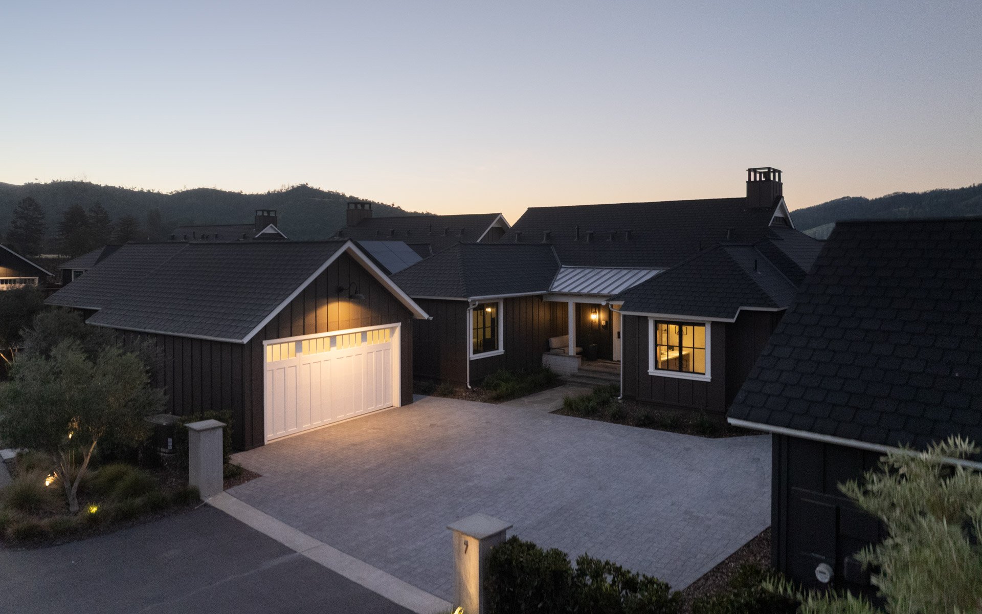 A suburban house at dusk with exterior lights on, black siding, a white garage door, and a paved driveway.